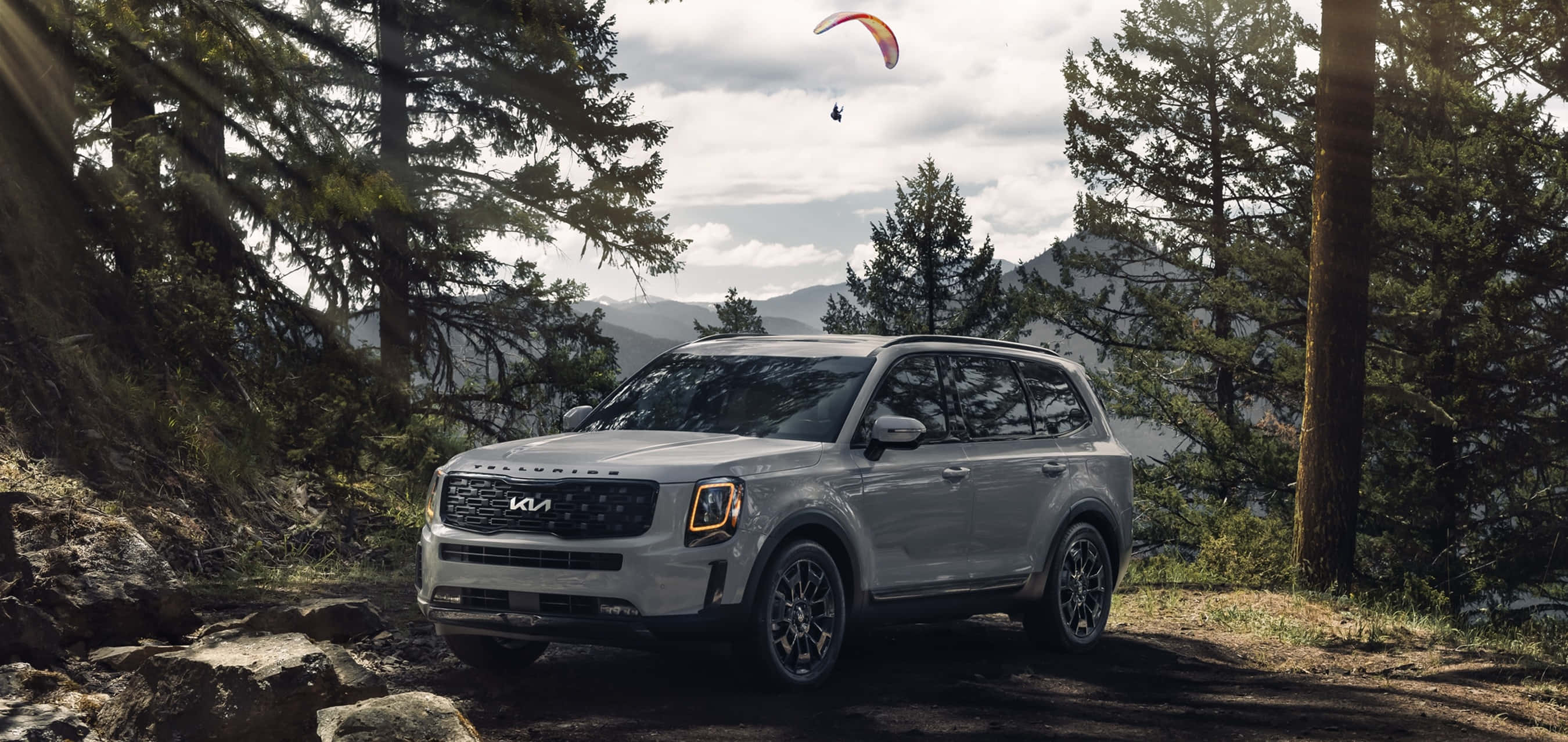 Kia Telluride 桌布 壁紙 — 一輛銀色Kia SUV停放在森林小徑上，滑翔傘在松樹和遠山上方翱翔。