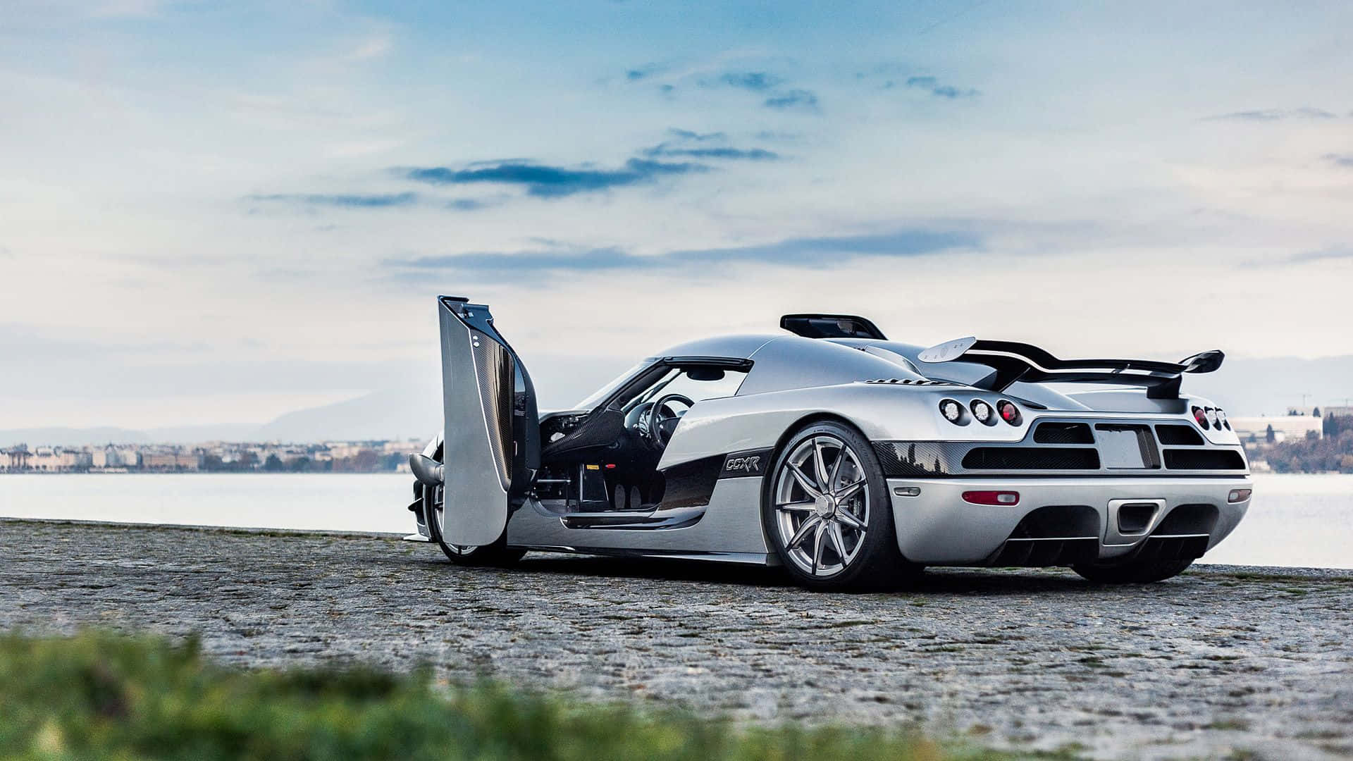 Koenigsegg 桌布 壁紙 — 一輛銀色Koenigsegg跑車打開車門，停泊在鵝卵石岸邊，旁邊是平靜的湖泊，天空部分多雲。