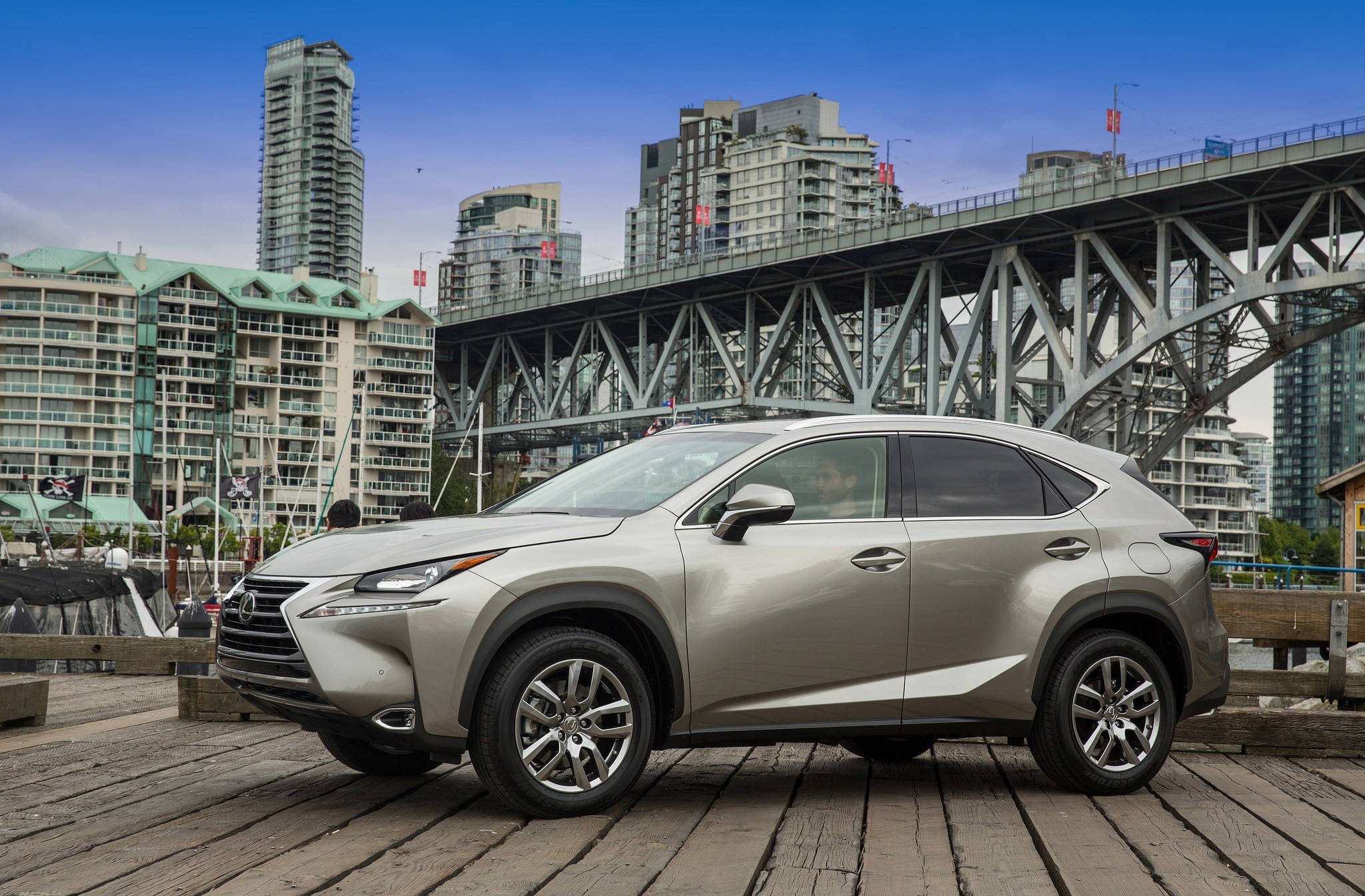 lexus iPhone 桌布 壁紙 — 一輛銀色Lexus NX SUV停泊在木製碼頭上，背景有鋼橋和現代城市天際線，天空清澈湛藍。