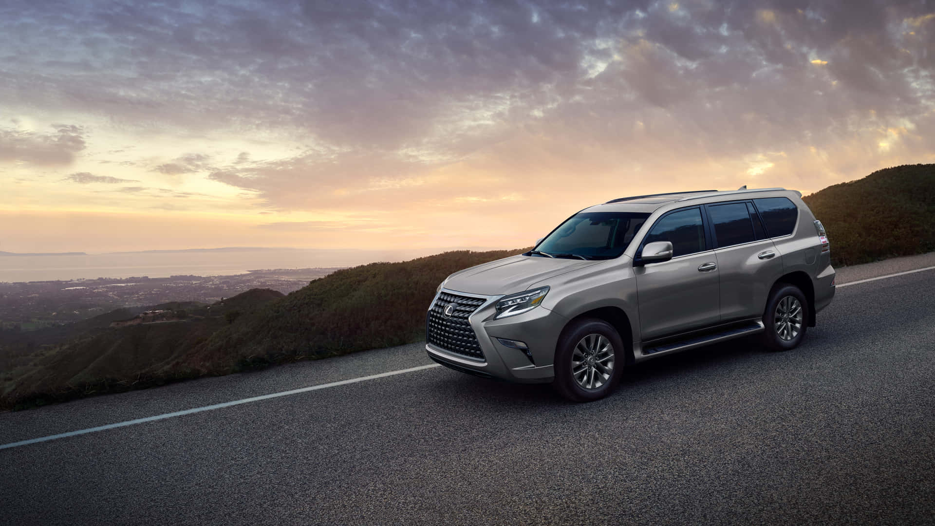 Lexus Gx 460 桌布 壁紙 — 一輛銀色Lexus SUV在日落時分沿著海岸山路行駛，天空佈滿戲劇性的雲層。