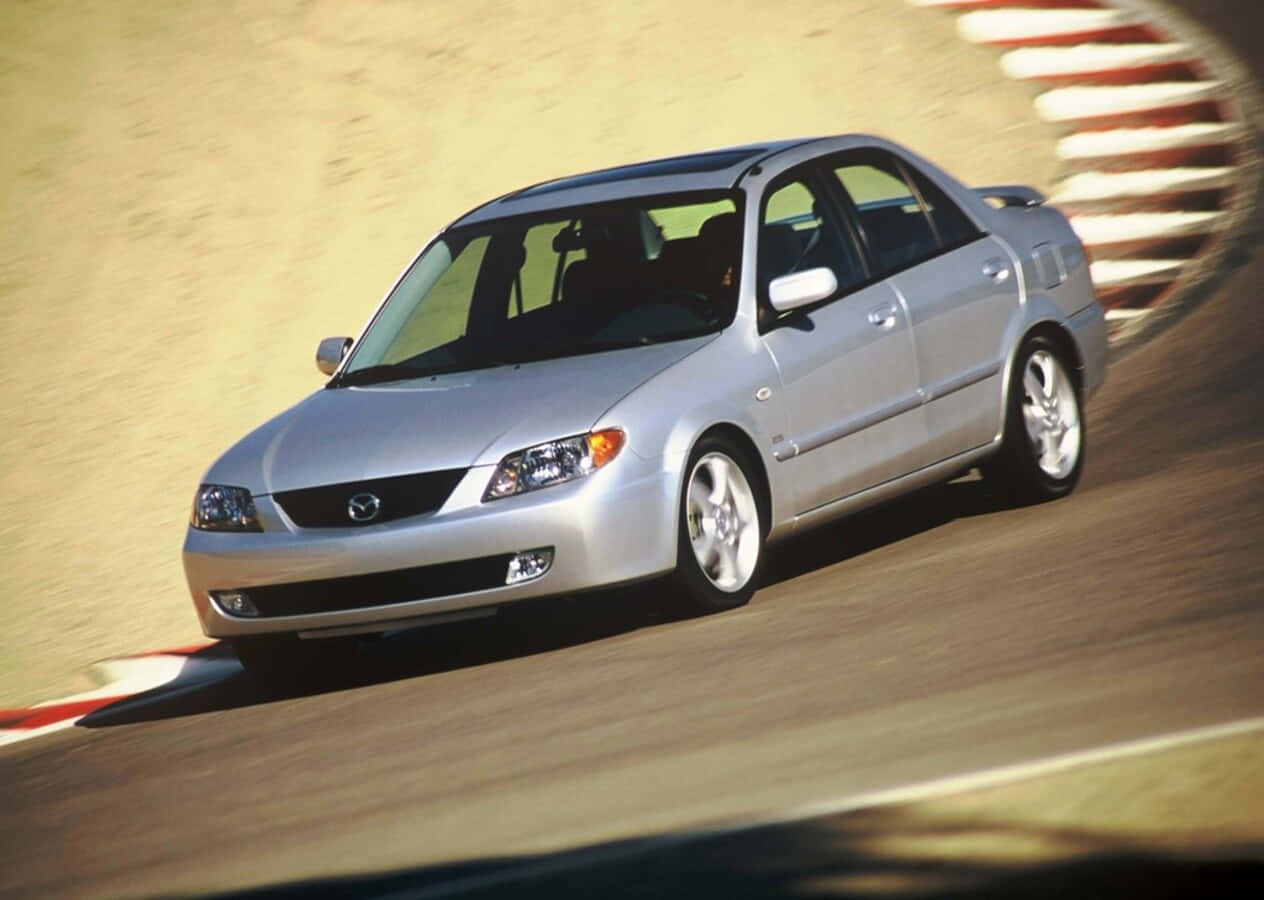 Mazda Protege 桌布 壁紙 — 一輛銀色Mazda房車高速駛過賽車場彎道，賽道表面呈現動態模糊效果。