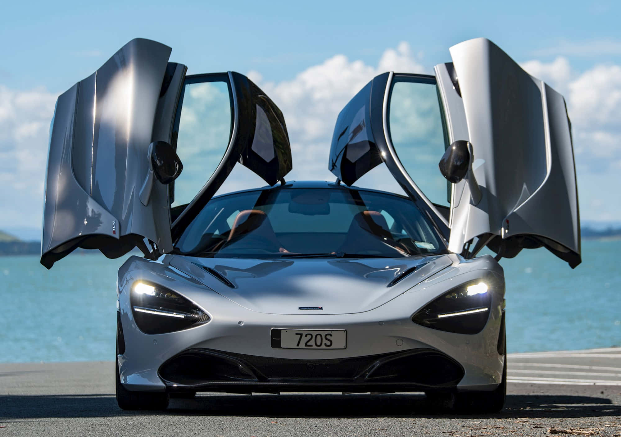 水 桌布 壁紙 — 一輛銀色McLaren 720S跑車，鷗翼車門打開，停在海岸公路上，天空湛藍。
