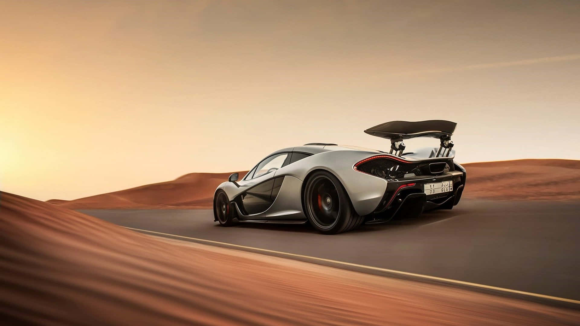麥拉倫 桌布 壁紙 — 一輛銀色McLaren P1超級跑車在日落時分沿沙漠公路飛馳，帶有大型後擾流板和模糊的背景。
