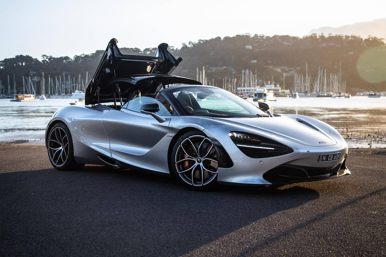 跑車 桌布 壁紙 — 一輛銀色McLaren跑車打開蝴蝶門，停泊在日落時分帆船附近的海濱道路上。