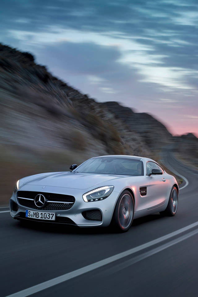 賓士 iPhone 桌布 壁紙 — 一輛銀色Mercedes-AMG GT在黃昏時分沿著蜿蜒的山路疾馳，背景是模糊的岩石地形。