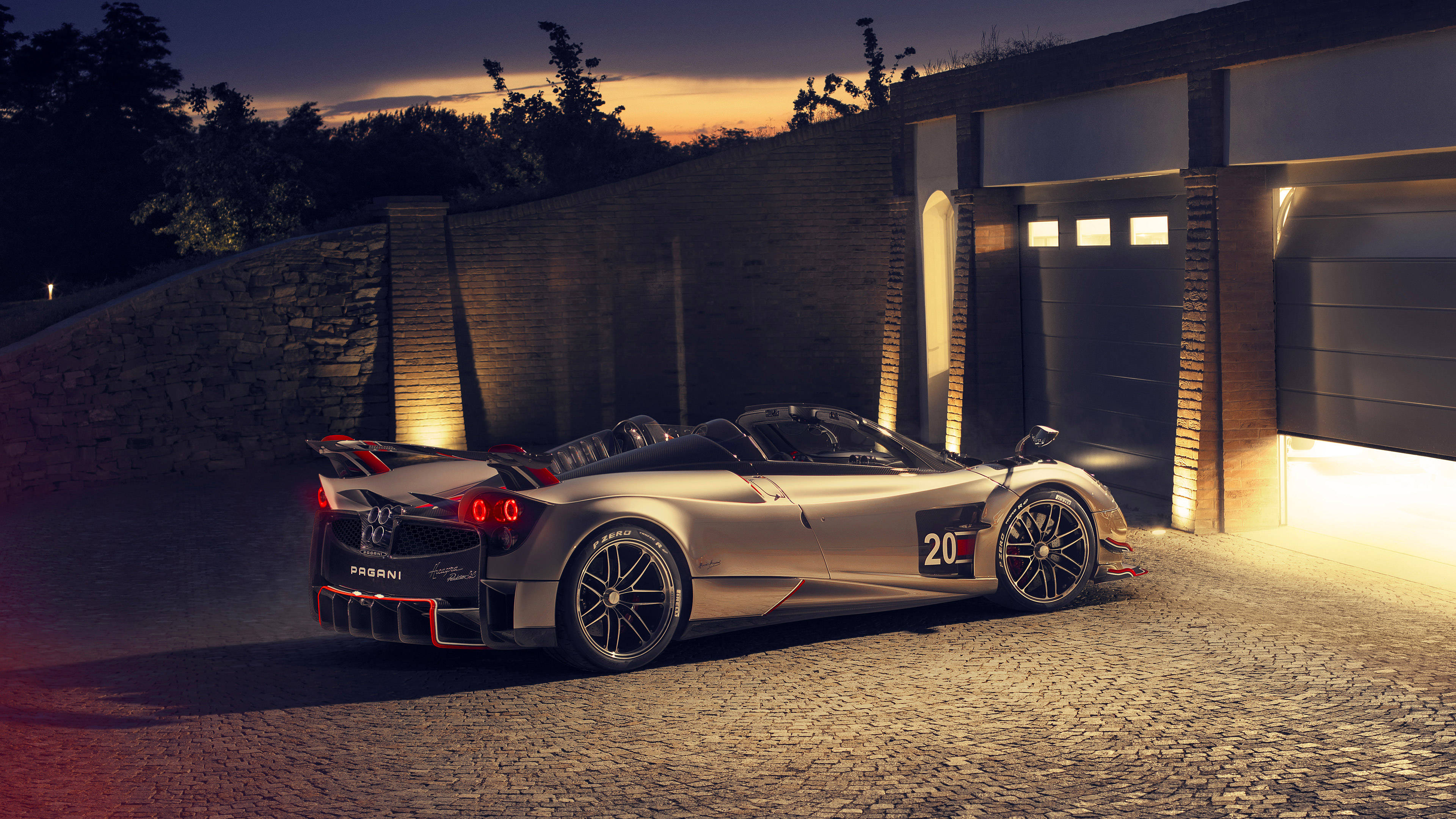 pagani iPhone 桌布 壁紙 — 一輛銀色Pagani Huayra Roadster在暮色燈光下閃閃發光，停在現代車庫旁的鵝卵石車道上。