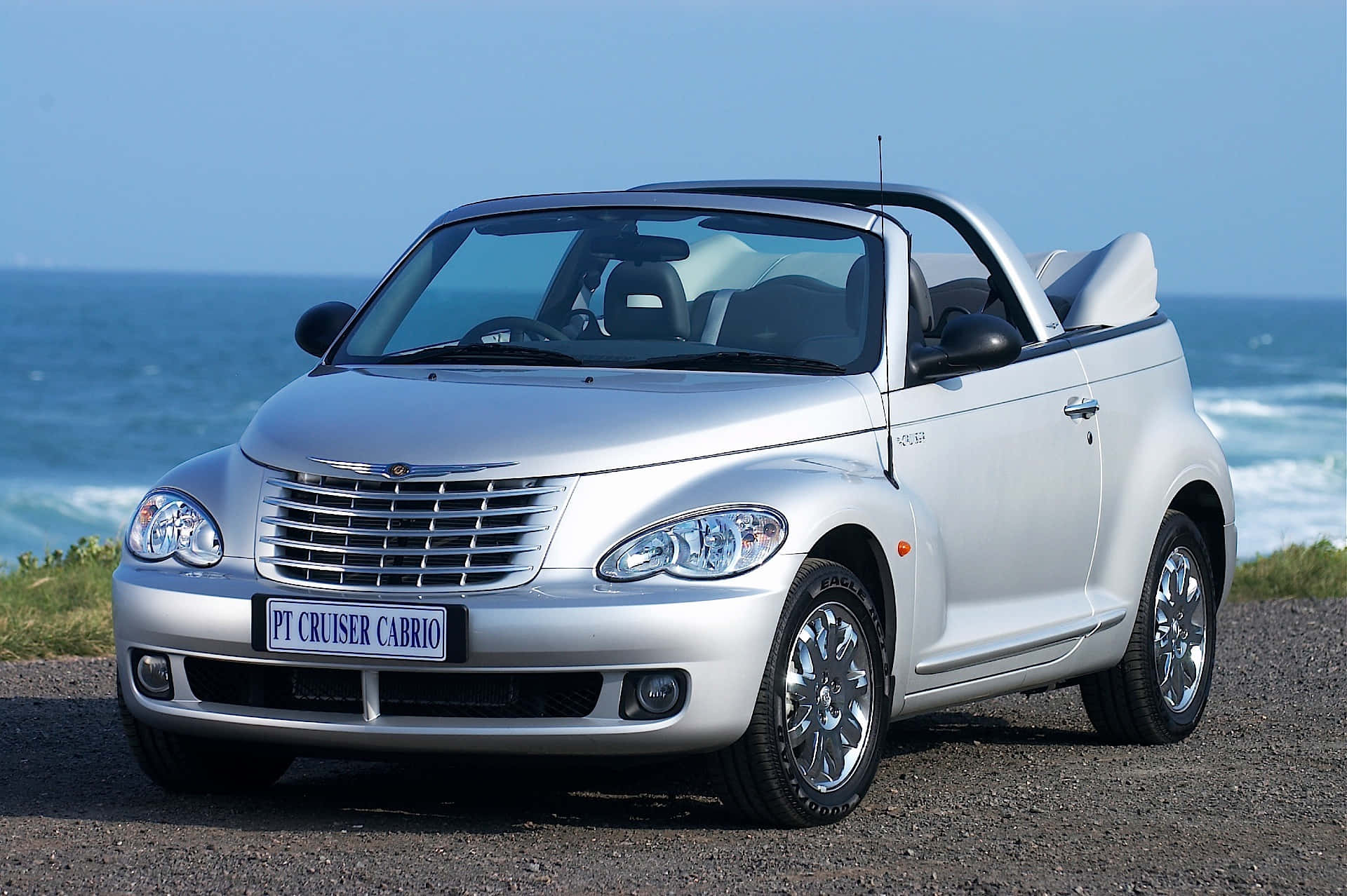 Chrysler Pt Cruiser 桌布 壁紙 — 一輛銀色PT Cruiser Cabrio敞篷車停泊在海岸碎石路上，背後是清澈藍天下拍岸的海浪。