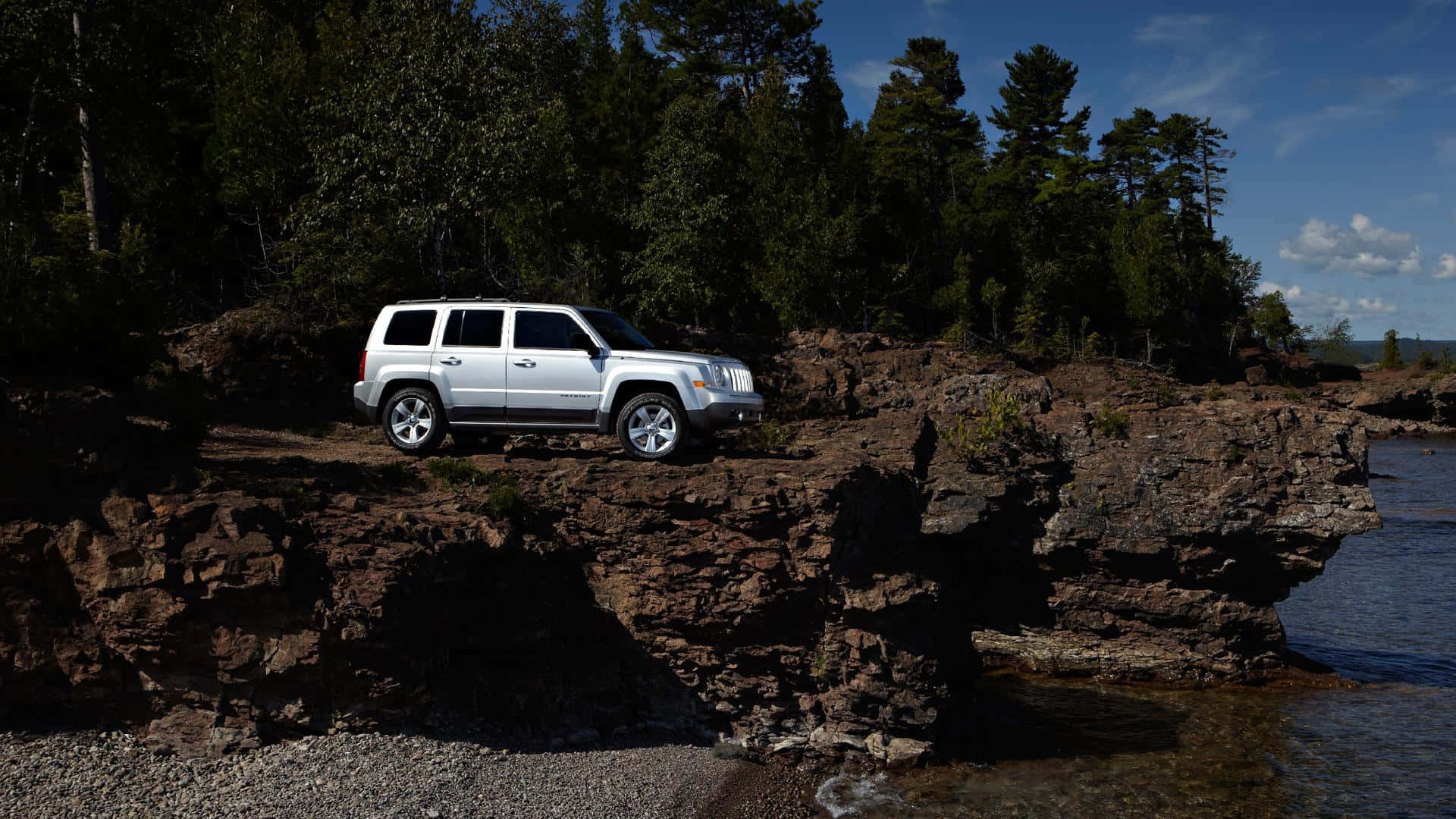 Jeep Patriot 桌布 壁紙 — 一輛銀色SUV停泊在崎嶇的懸崖上，俯瞰著被茂密常綠樹木環繞的平靜湖泊，上方是明亮的藍天。