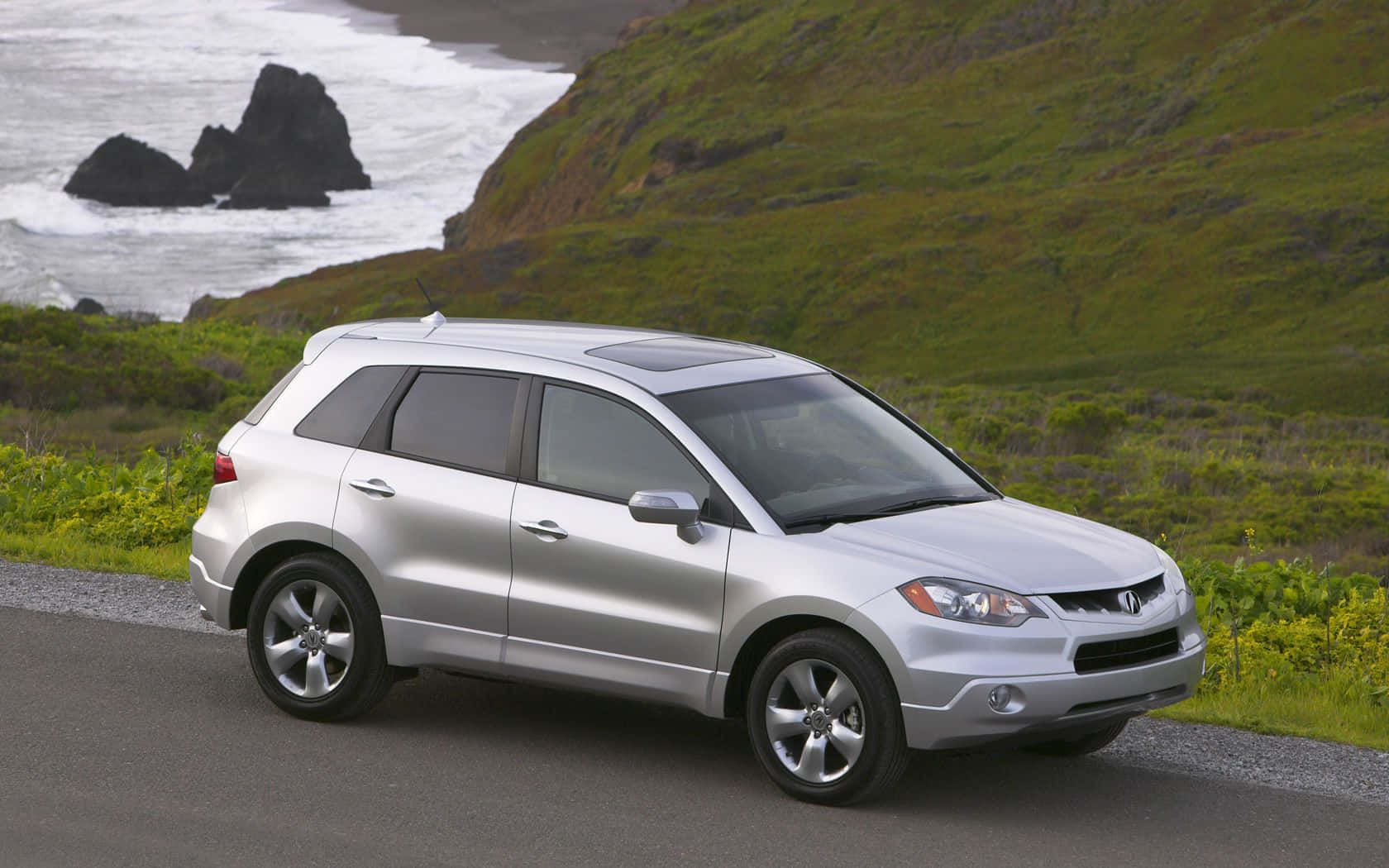 Acura Rdx 桌布 壁紙 — 一輛銀色SUV停泊在沿海公路上，背景有綠色山丘和拍打的海浪。