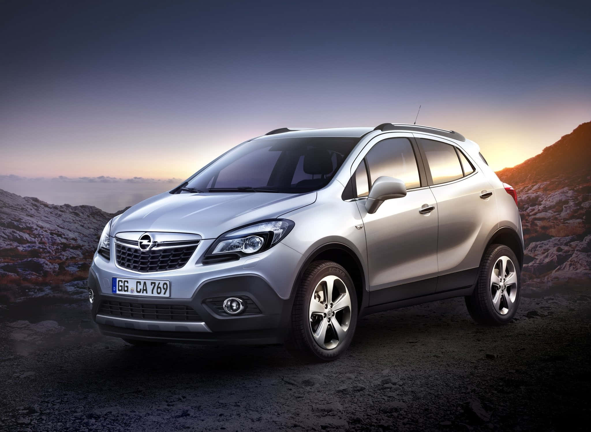 Opel Mokka 桌布 壁紙 — 一輛銀色SUV在日落時分停泊在崎嶇的山路上，溫暖的光芒在岩石山丘後閃耀。