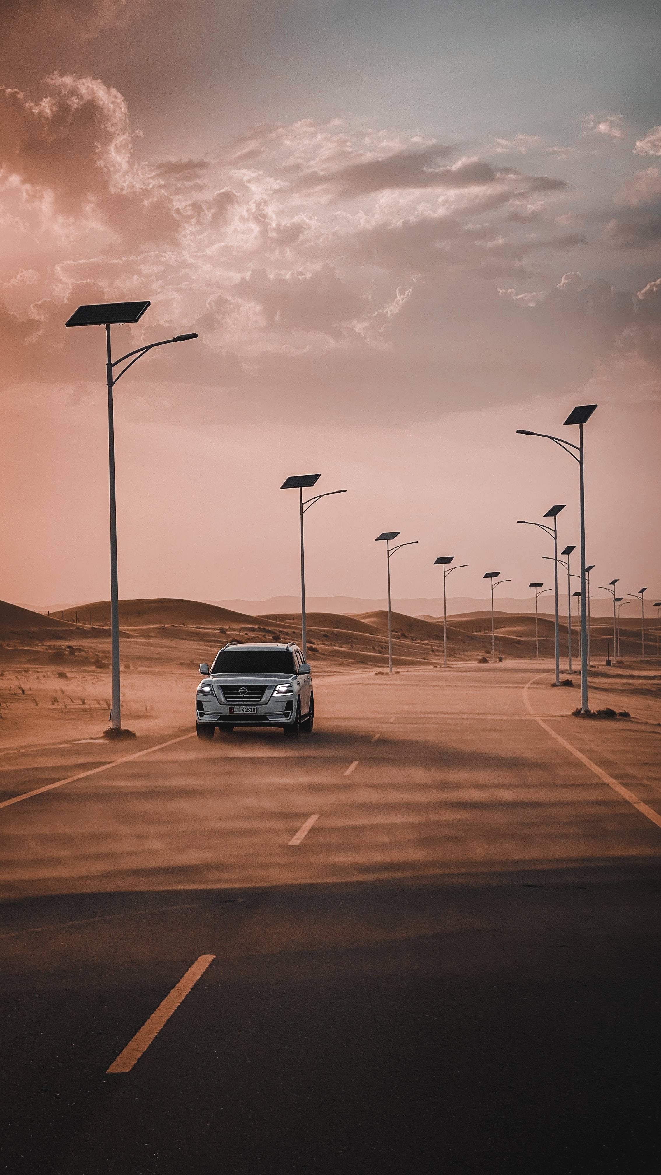 Nissan 桌布 壁紙 — 一輛銀色SUV在溫暖的日落天空下行駛在一條長長的沙漠公路上，公路兩旁是太陽能路燈。