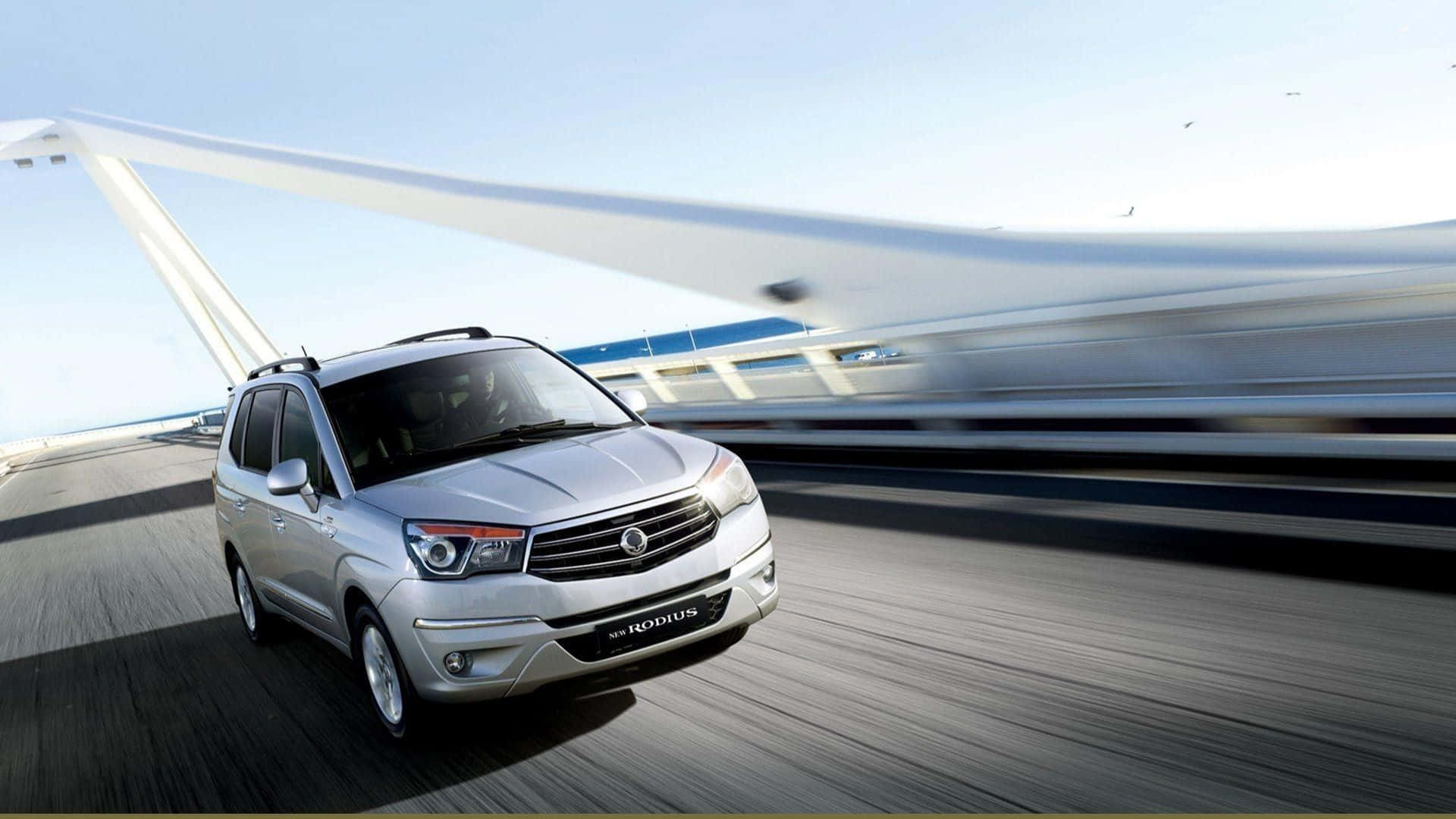 Ssangyong 桌布 壁紙 — 一輛銀色SUV在現代橋樑上疾馳，動態模糊效果強調了晴朗天空下的速度。