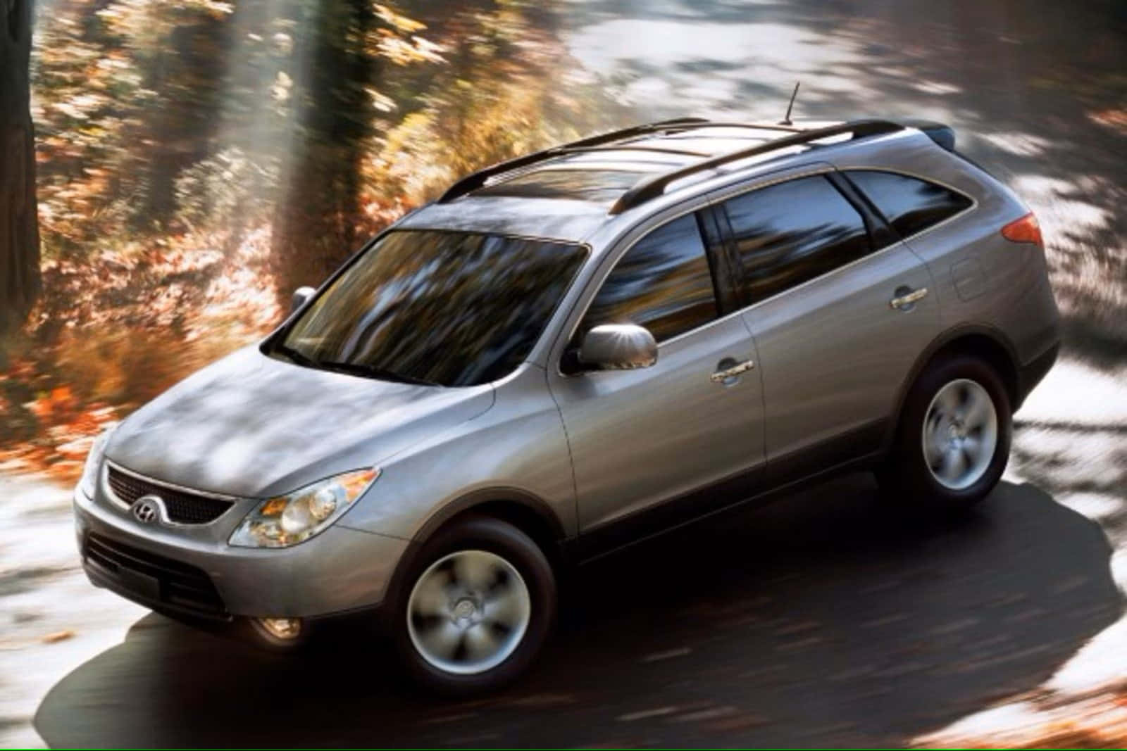 Hyundai Veracruz 桌布 壁紙 — 一輛銀色SUV在陽光明媚的秋日森林中行駛，帶有模糊的動態和飄落的樹葉。
