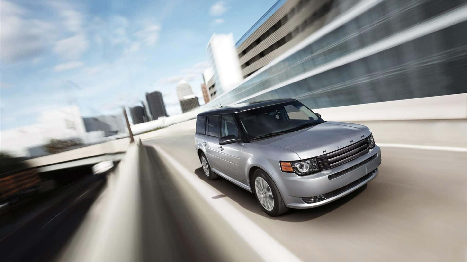 Ford Flex 桌布 壁紙 — 一輛銀色SUV在高速公路上疾馳，模糊的城市建築在明亮的藍天下掠過。