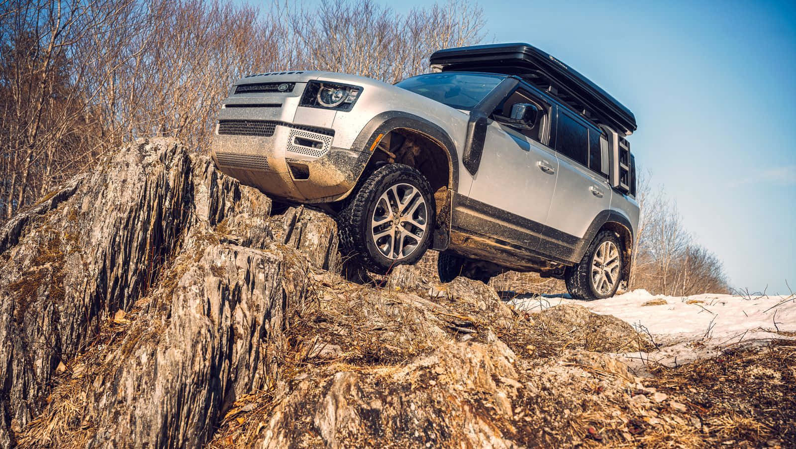 Land Rover Defender 桌布 壁紙 — 一輛銀色SUV配備車頂行李架，在晴朗的藍天下征服崎嶇的雪岩斜坡。