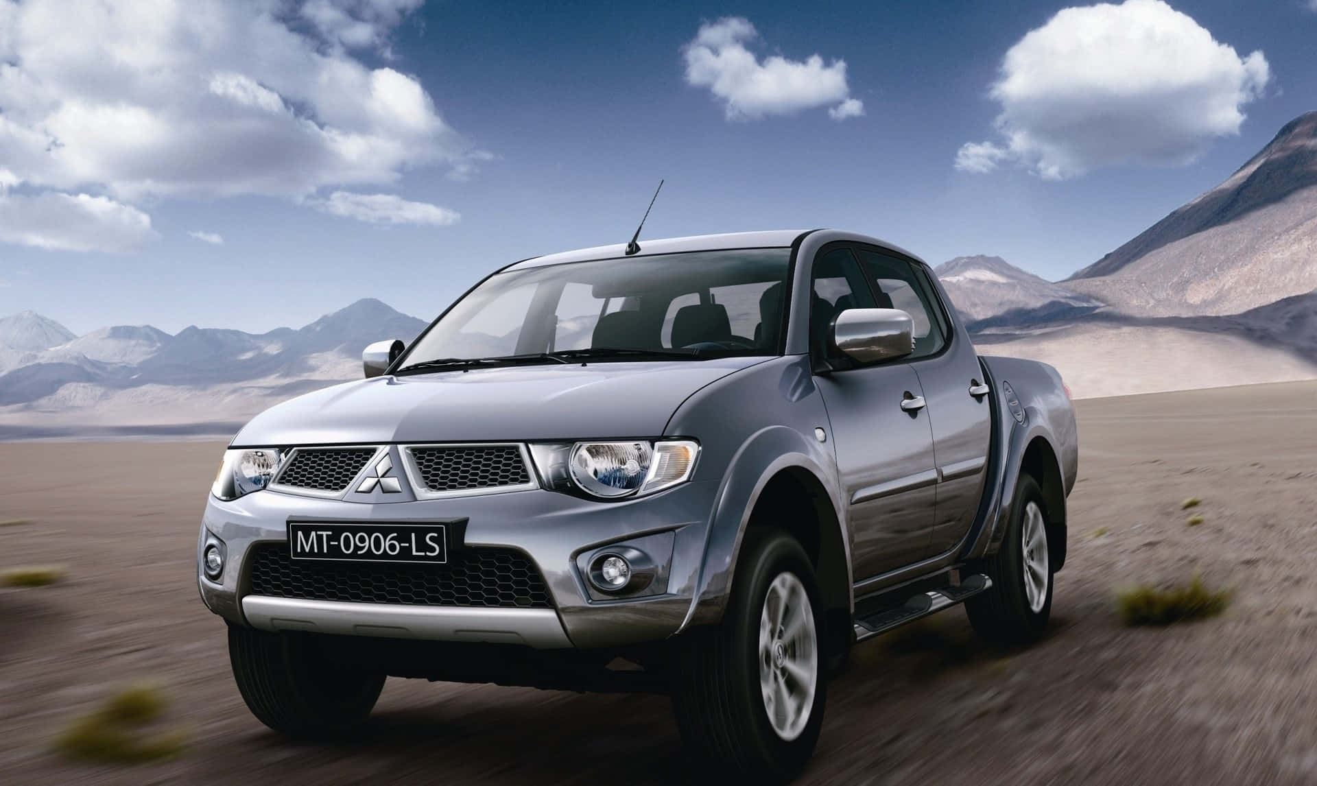 Mitsubishi L200 桌布 壁紙 — 一輛銀色三菱皮卡車在塵土飛揚的沙漠景觀中疾馳，背景有山脈和零星雲朵。