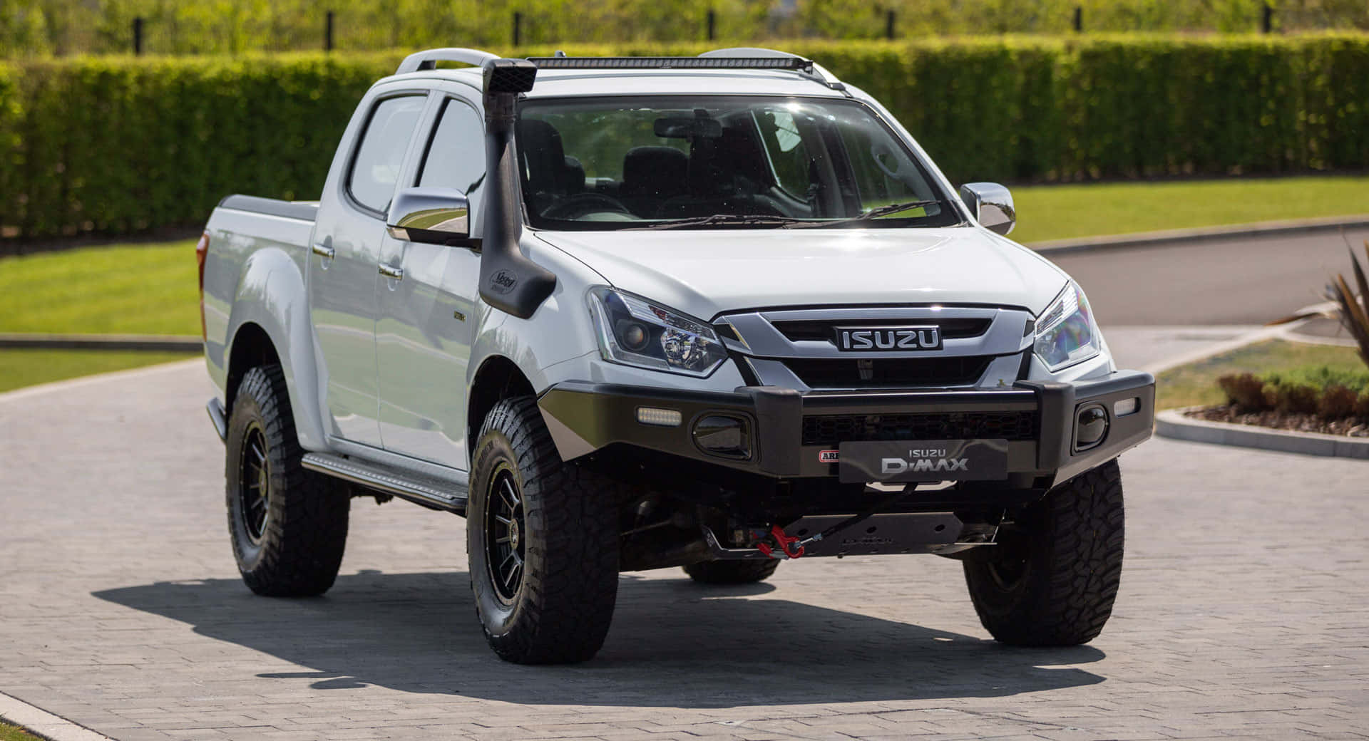 Isuzu 桌布 壁紙 — 一輛銀色五十鈴D-Max皮卡車，配備黑色越野輪胎和前保險桿，停在鋪砌的車道上，周圍環繞著綠色籬笆和草地。