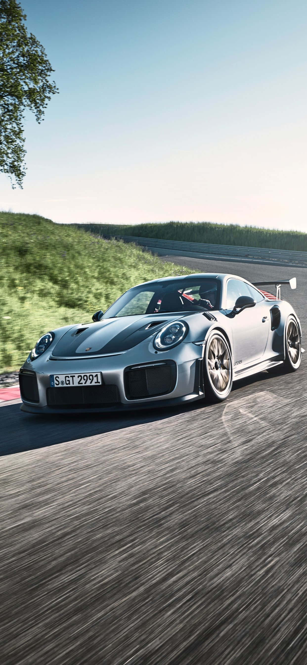 Porsche iPhone 桌布 壁紙 — 一輛銀色保時捷 911 GT2 RS 在蜿蜒的賽道上高速行駛，帶有動態模糊效果，背景是綠色的山丘和清澈的藍天。