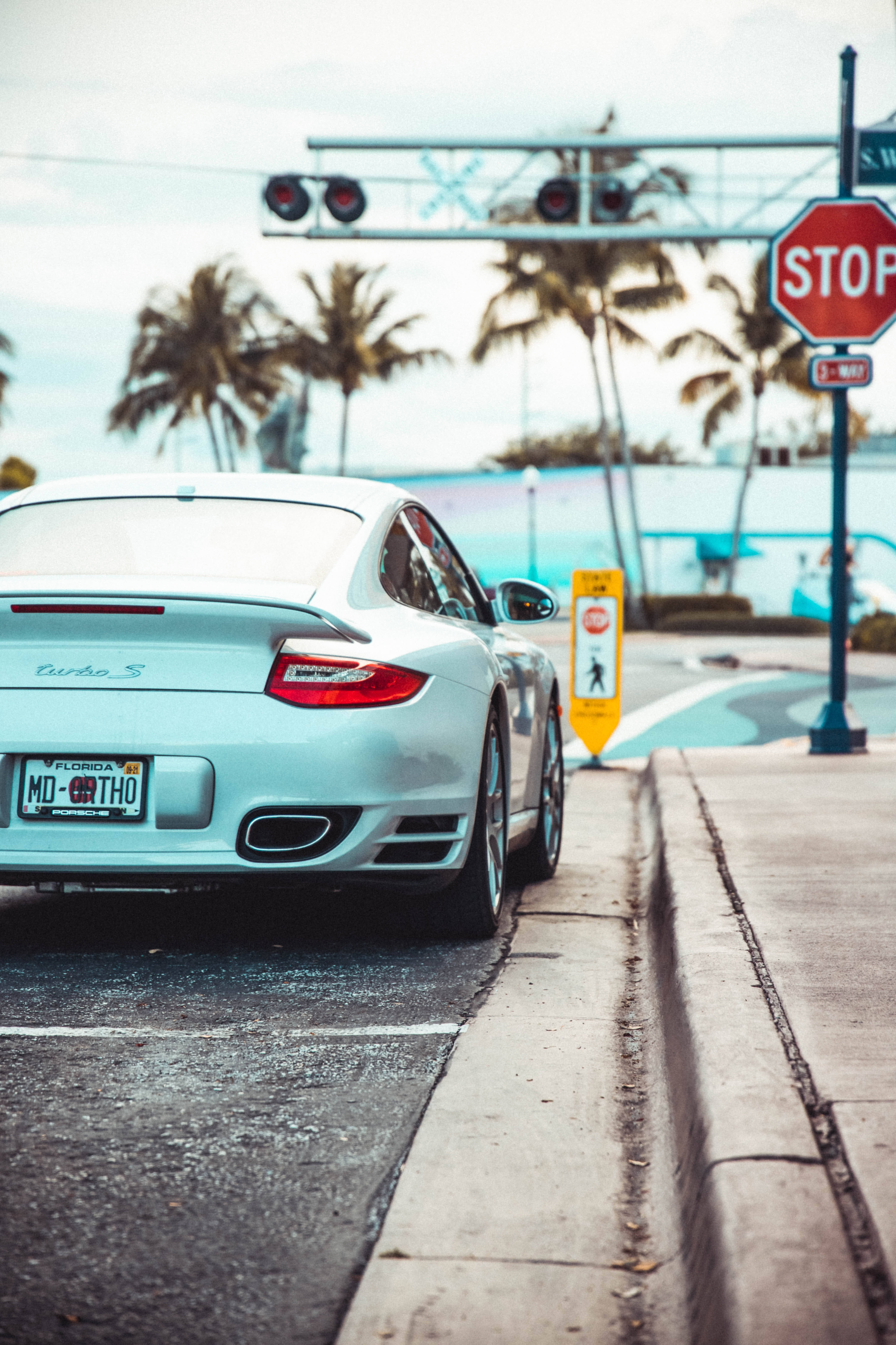 保時捷 桌布 壁紙 — 一輛銀色保時捷 Turbo S 停在停車標誌處，背景有棕櫚樹和海洋，天空多雲。