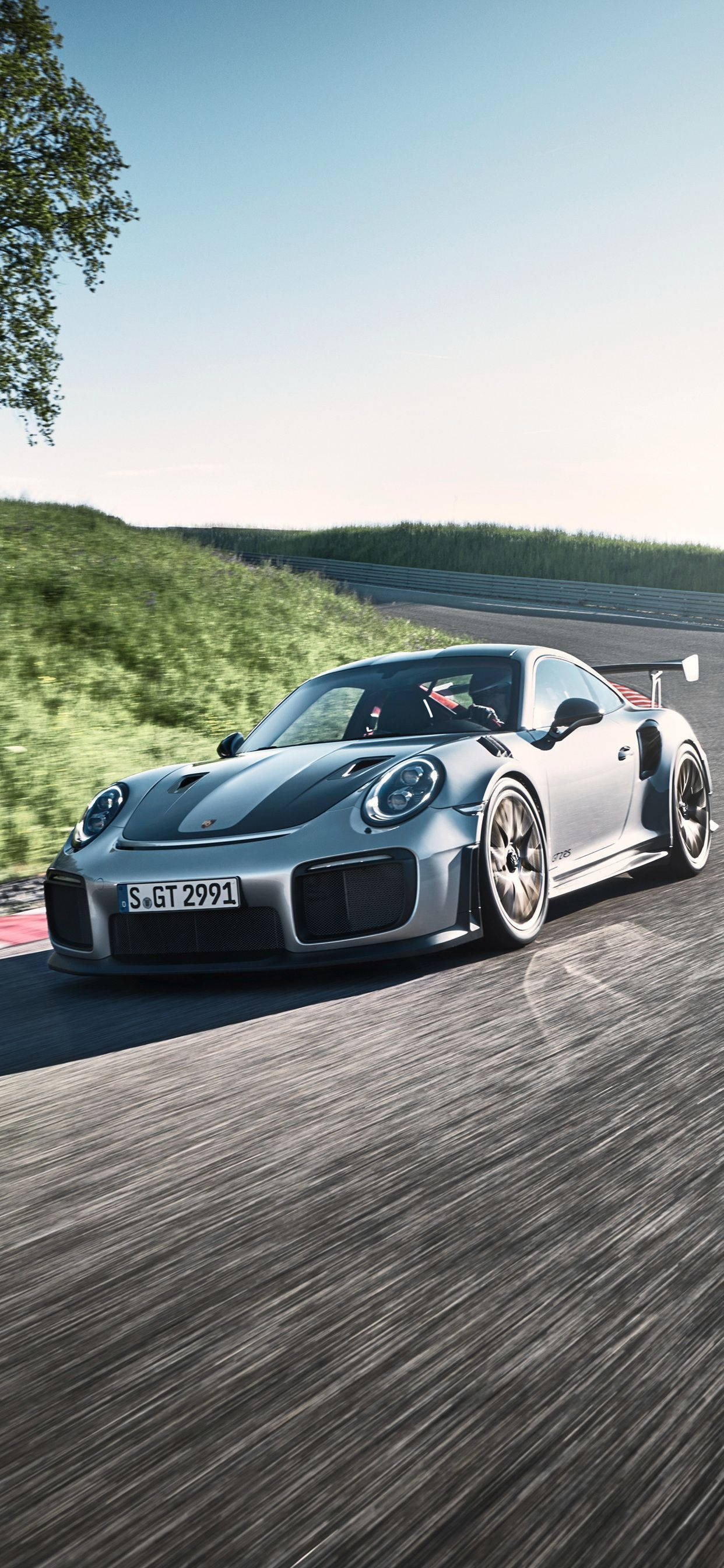 保時捷 911 桌布 壁紙 — 一輛銀色保時捷911 GT2 RS在蜿蜒的賽道上疾馳，帶有動態模糊效果，背景是綠色的山丘和清澈的藍天。