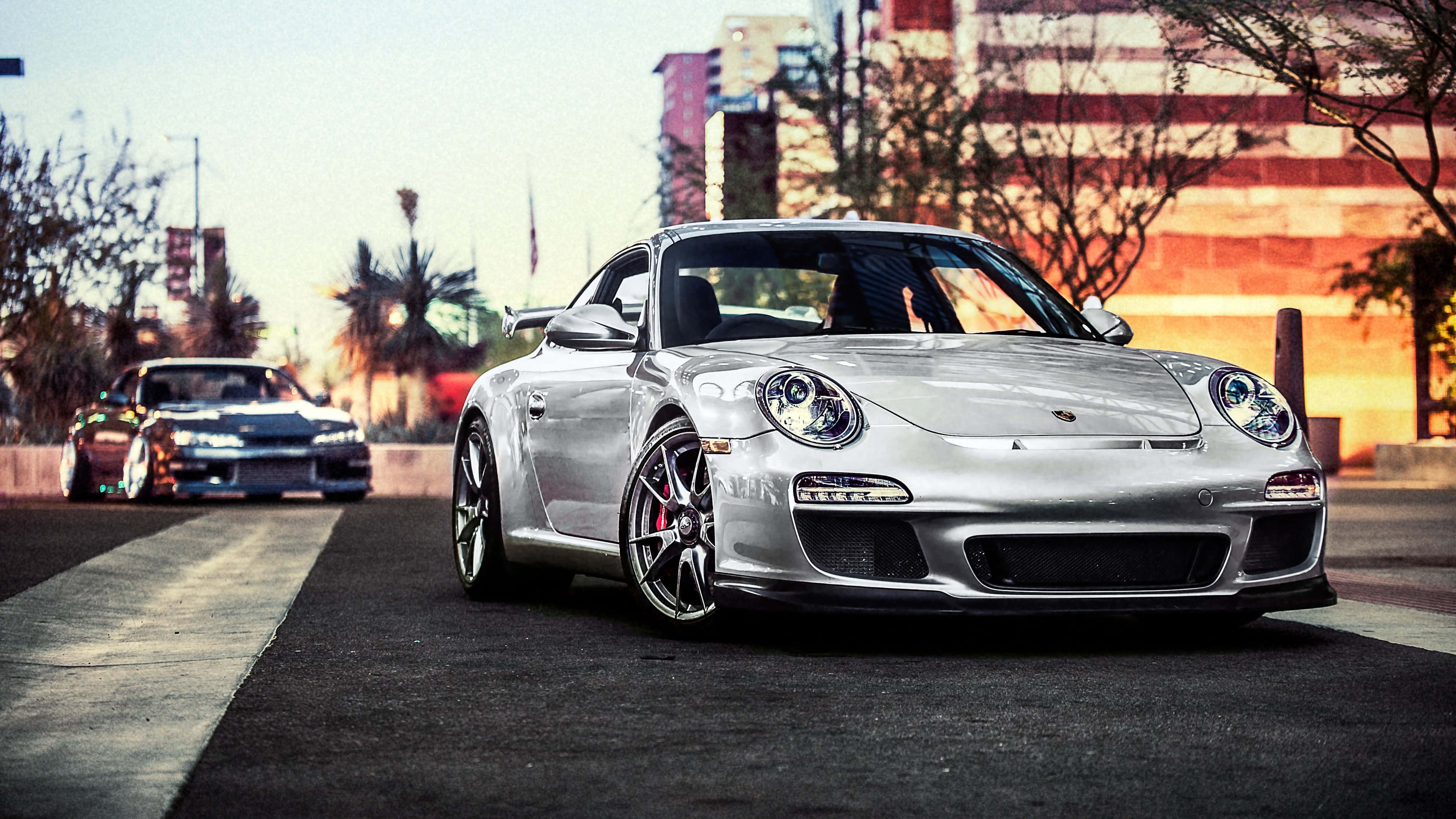 桌布 壁紙 — 一輛銀色保時捷911 GT3 RS停泊在日落時分的城市街道上，背景有棕櫚樹和建築物。