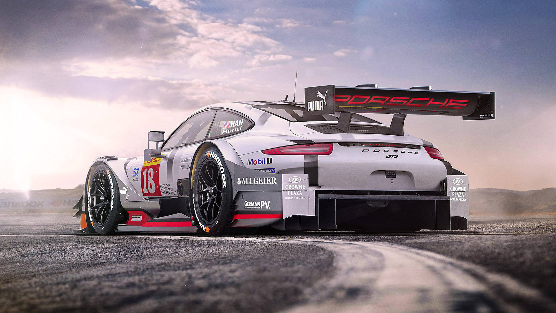 Auto Racing 桌布 壁紙 — 一輛銀色保時捷911 GT3 R賽車，編號18，在戲劇性的日出天空下，沿著蜿蜒的瀝青賽道飛馳。