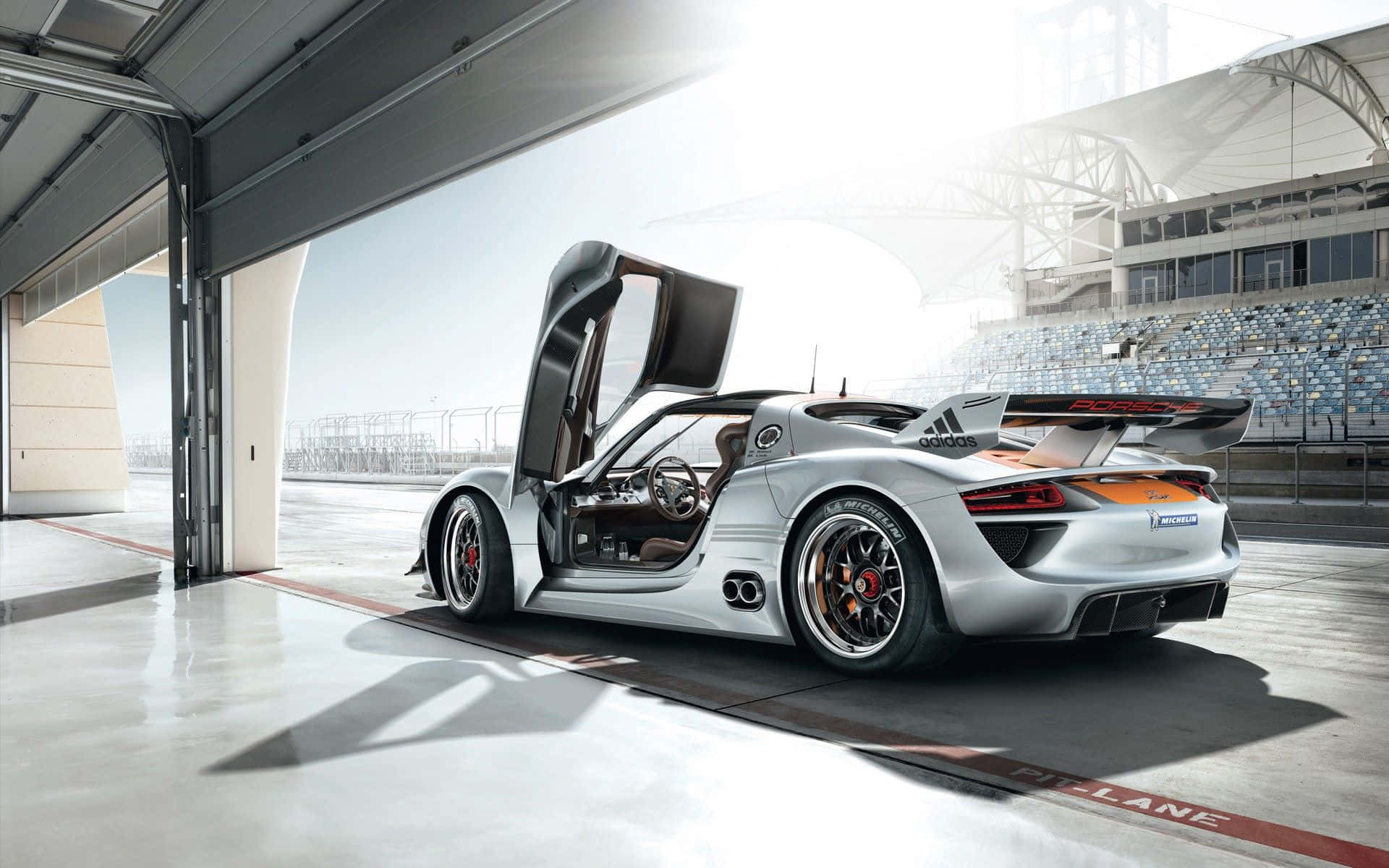 Hybrid Car 桌布 壁紙 — 一輛銀色保時捷918 Spyder，鷗翼車門敞開，停在賽車庫內，面向陽光普照的賽道。