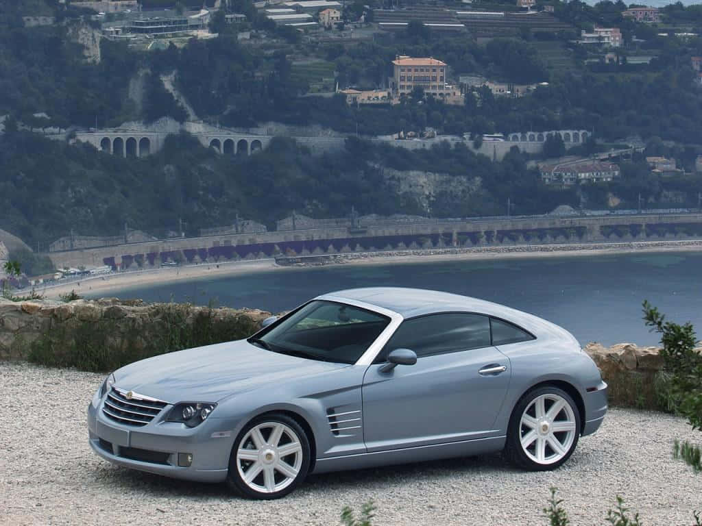 Chrysler Crossfire 桌布 壁紙 — 一輛銀色克萊斯勒Crossfire跑車停泊在碎石觀景點，背景有沿海小鎮和拱橋。