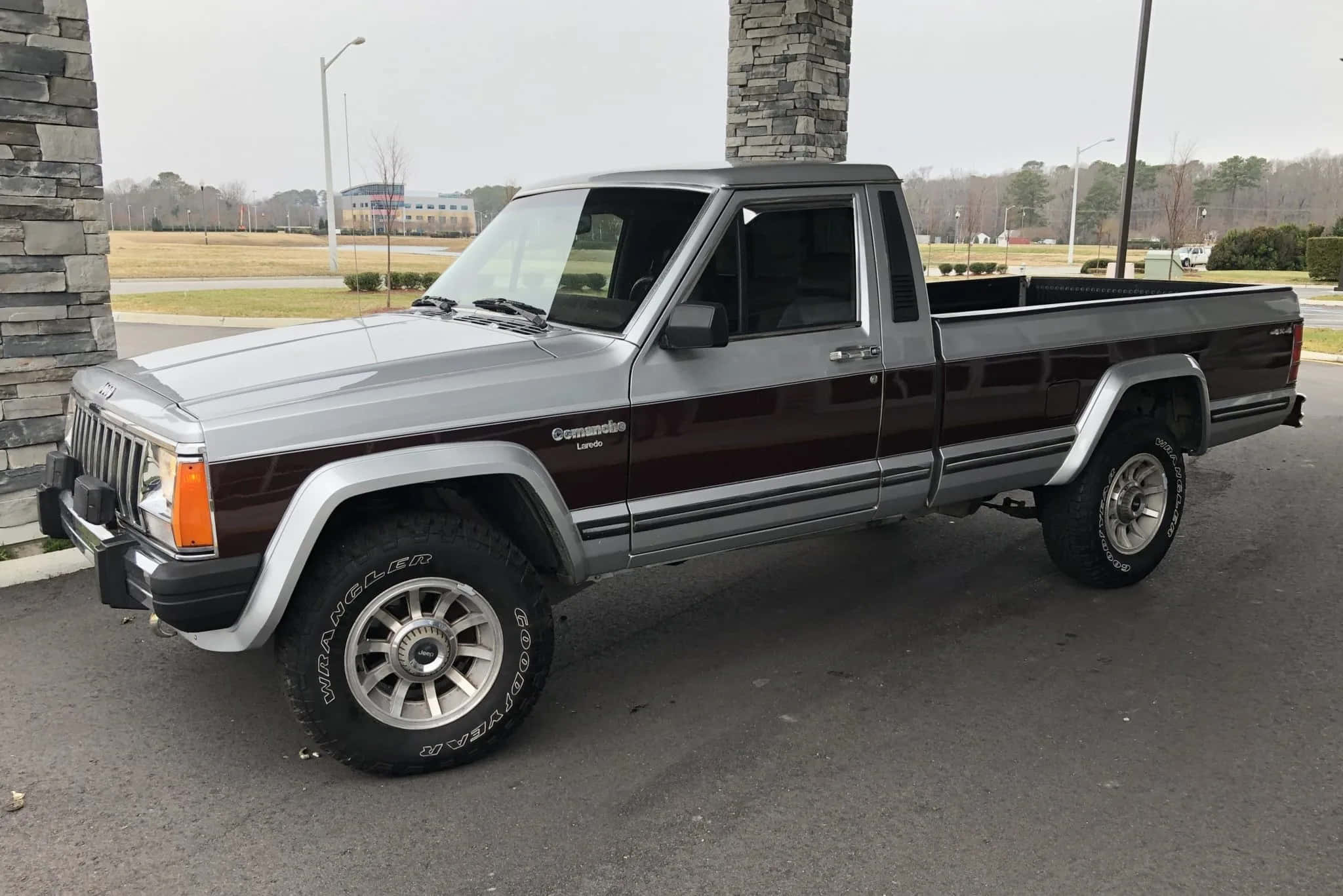 Jeep Comanche 桌布 壁紙 — 一輛銀色和栗色的Jeep Comanche pickup貨車停泊在瀝青路上，旁邊是石柱，天空多雲。
