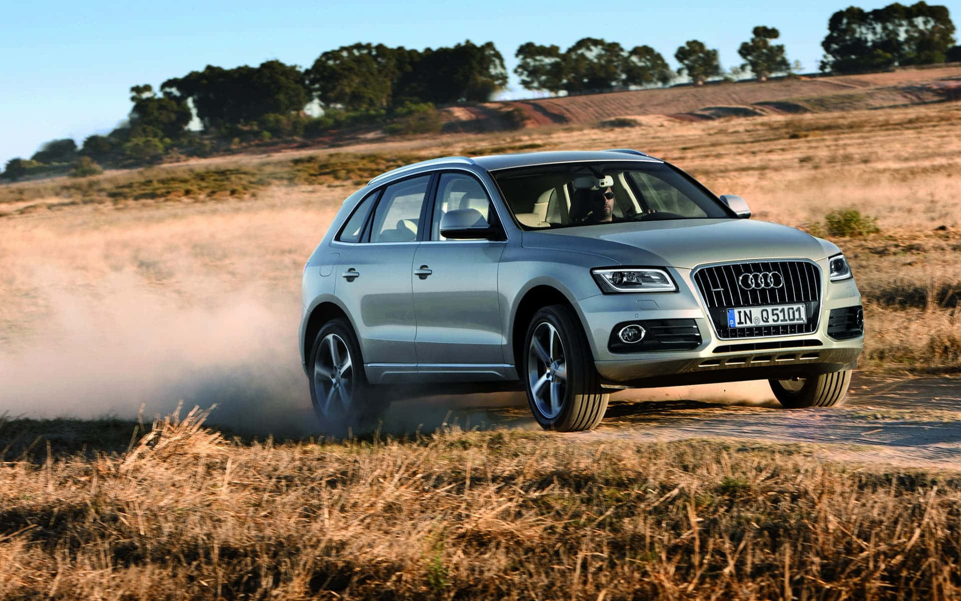Audi Sq5 桌布 壁紙 — 一輛銀色奧迪Q5 SUV在晴朗藍天下駛過乾燥草叢中的泥路，揚起塵土。