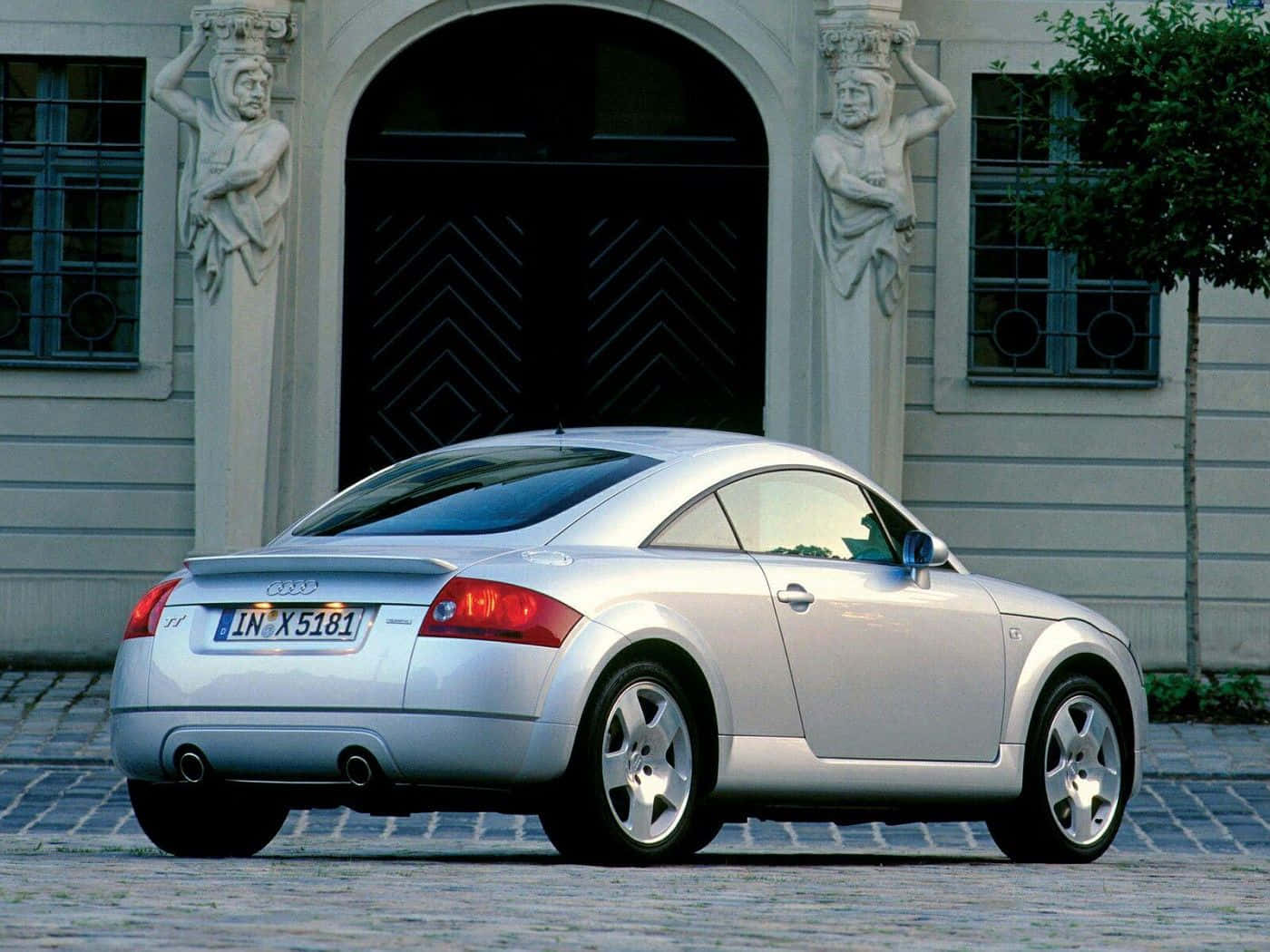 Audi Tt 桌布 壁紙 — 一輛銀色奧迪TT雙門跑車停放在鵝卵石庭院，前方是一座宏偉建築，飾有華麗的石像。