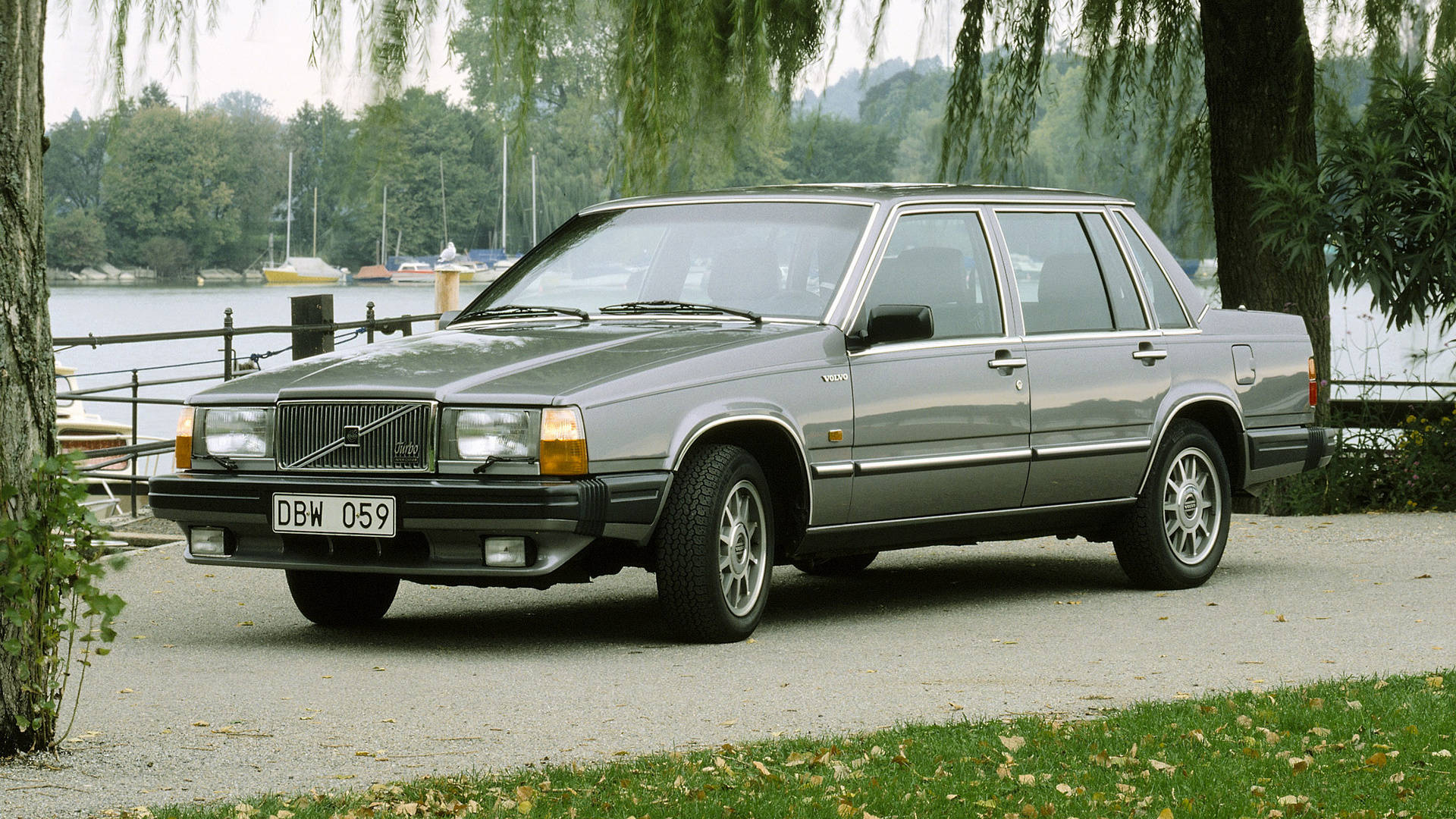 volvo iPhone 桌布 壁紙 — 一輛銀色富豪240房車停泊在湖邊的碎石路上，背景有垂柳和帆船。