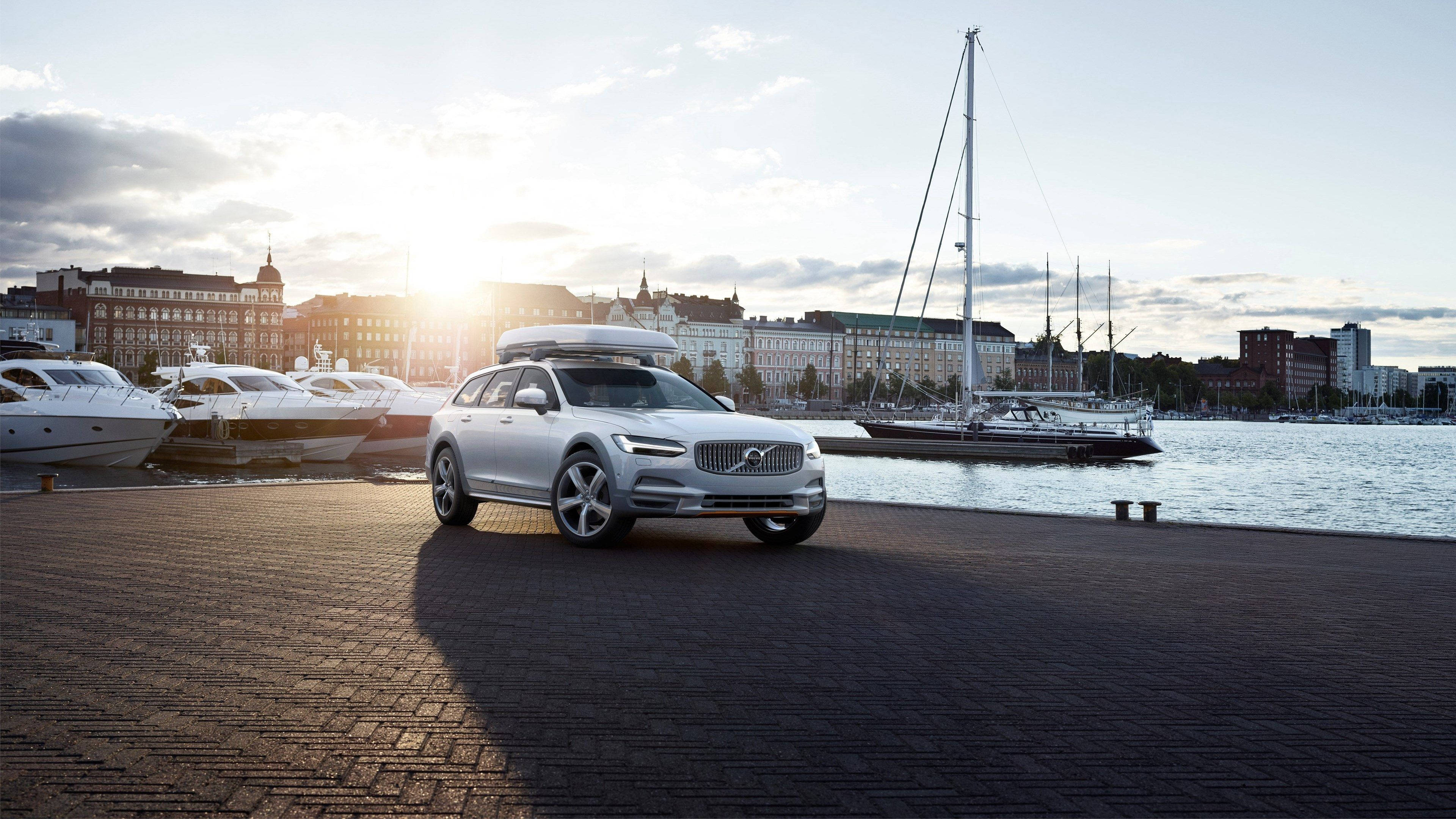 Volvo 桌布 壁紙 — 一輛銀色富豪旅行車連車頂箱停泊在磚碼頭上，夕陽西下，旁邊有帆船和城市建築。