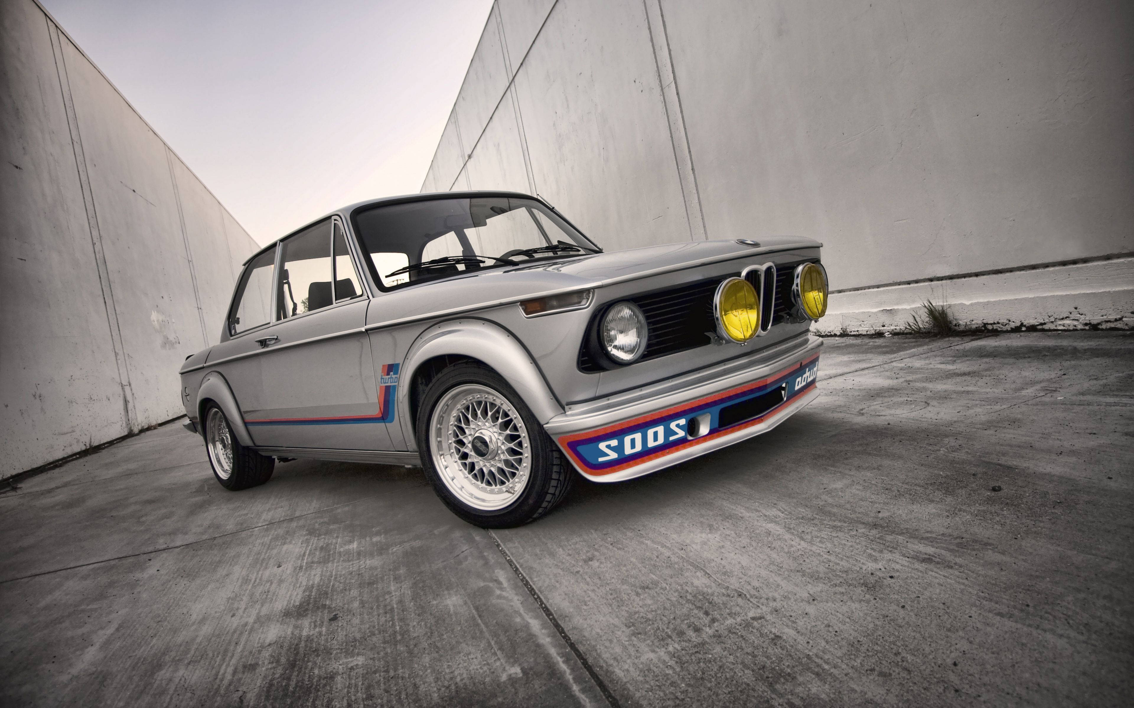 classic bmw 桌布 壁紙 — 一輛銀色寶馬賽車，黃色頭燈，藍紅條紋，停在混凝土隧道中。