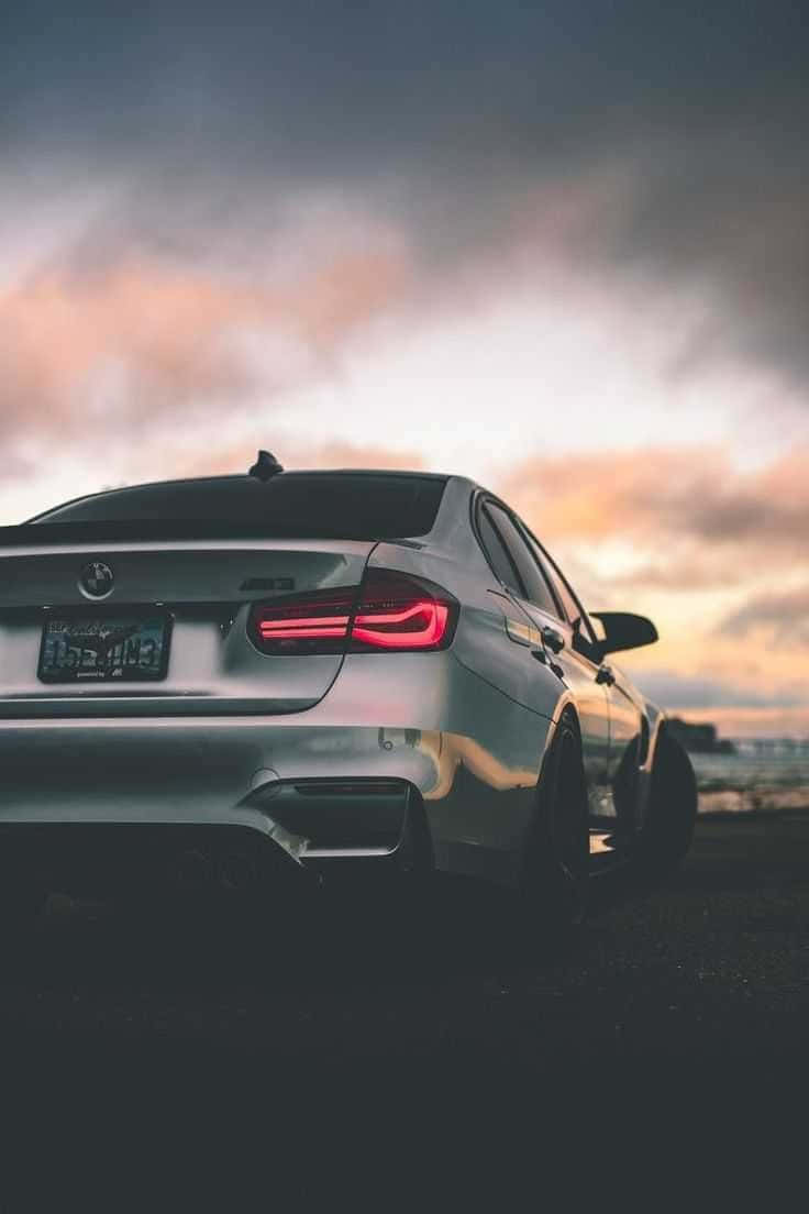 Bmw iPhone 桌布 壁紙 — 一輛銀色寶馬轎車在戲劇性的暮色天空下停在黑暗的海岸上，紅色尾燈閃閃發光。