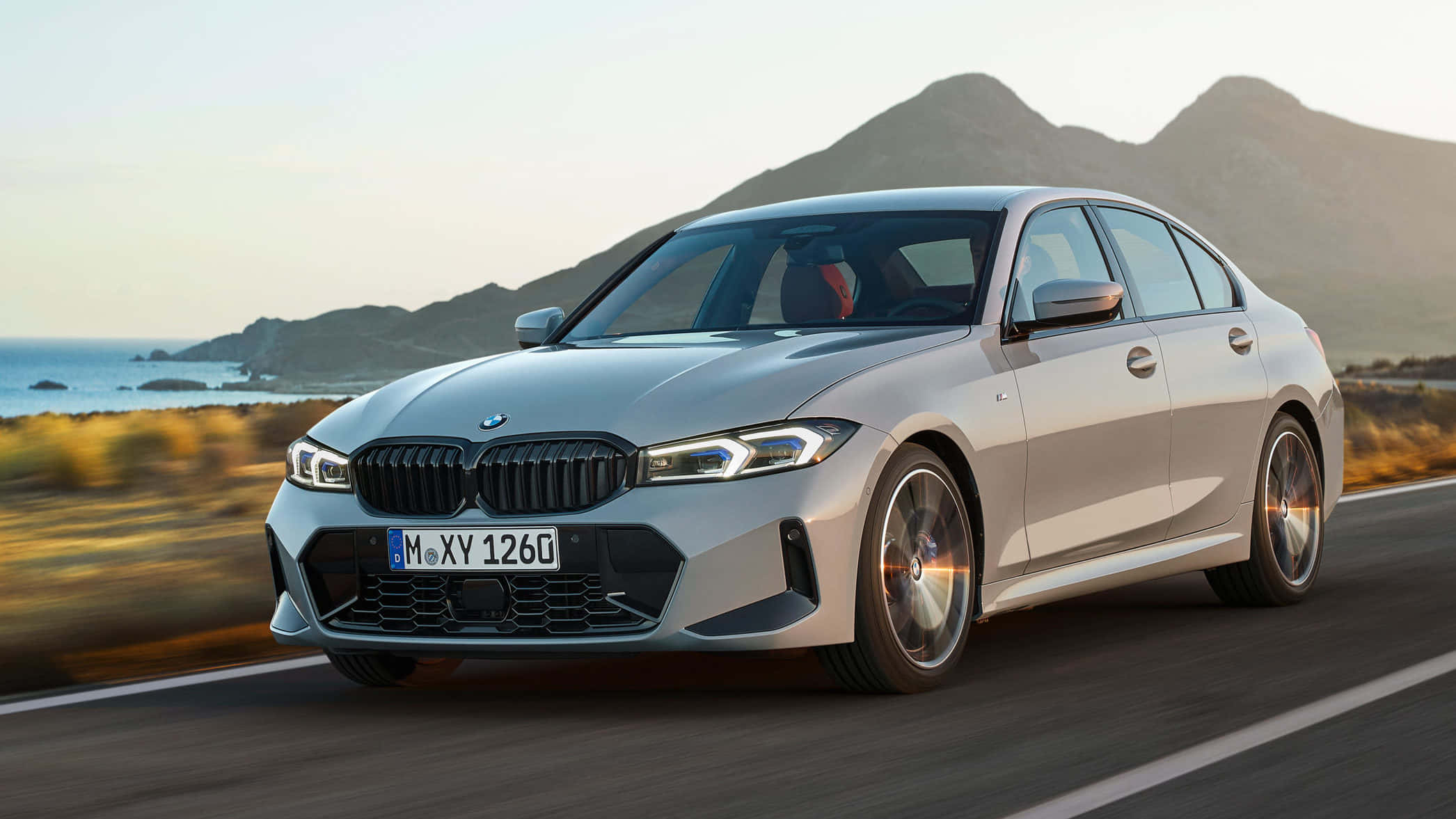 Bmw 3 Series 桌布 壁紙 — 一輛銀色寶馬轎車在黃金時段沿著海岸公路疾駛，背景是山脈和海洋。