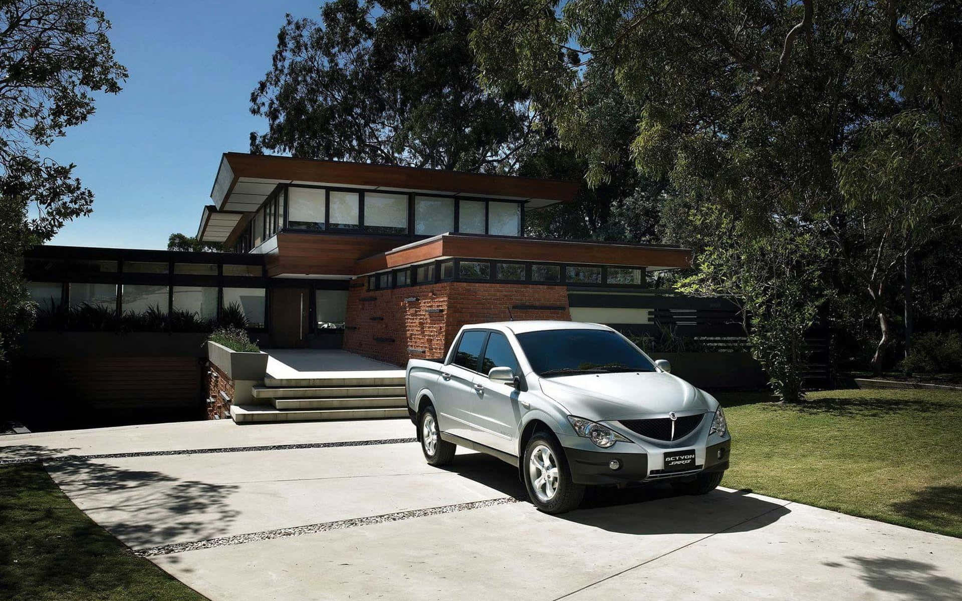 Ssangyong 桌布 壁紙 — 一輛銀色小型貨車停放在現代木磚房屋前的混凝土車道上，周圍綠樹成蔭，天空湛藍。