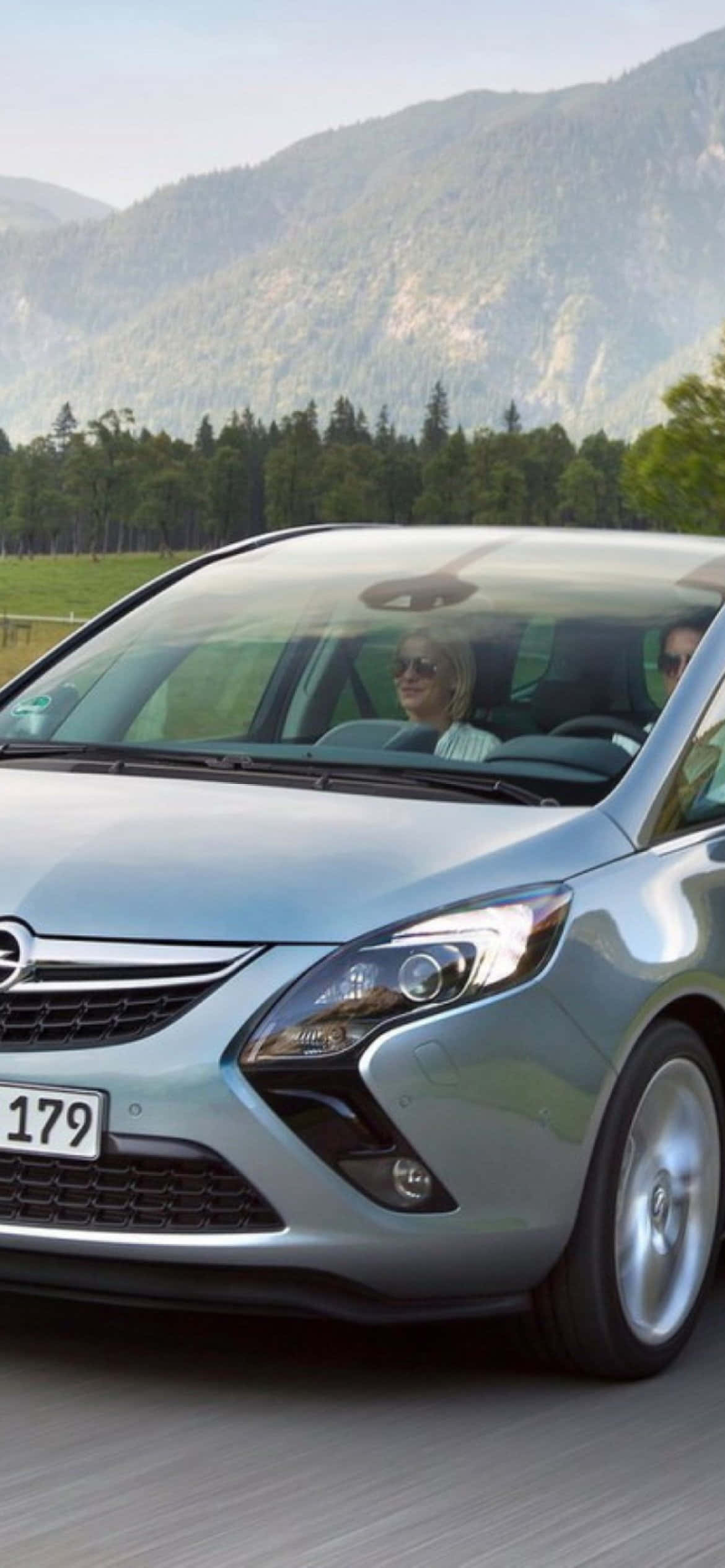 Opel Zafira 桌布 壁紙 — 一輛銀色小型貨車在明亮的天空下，沿著一條有森林覆蓋山脈的道路行駛。