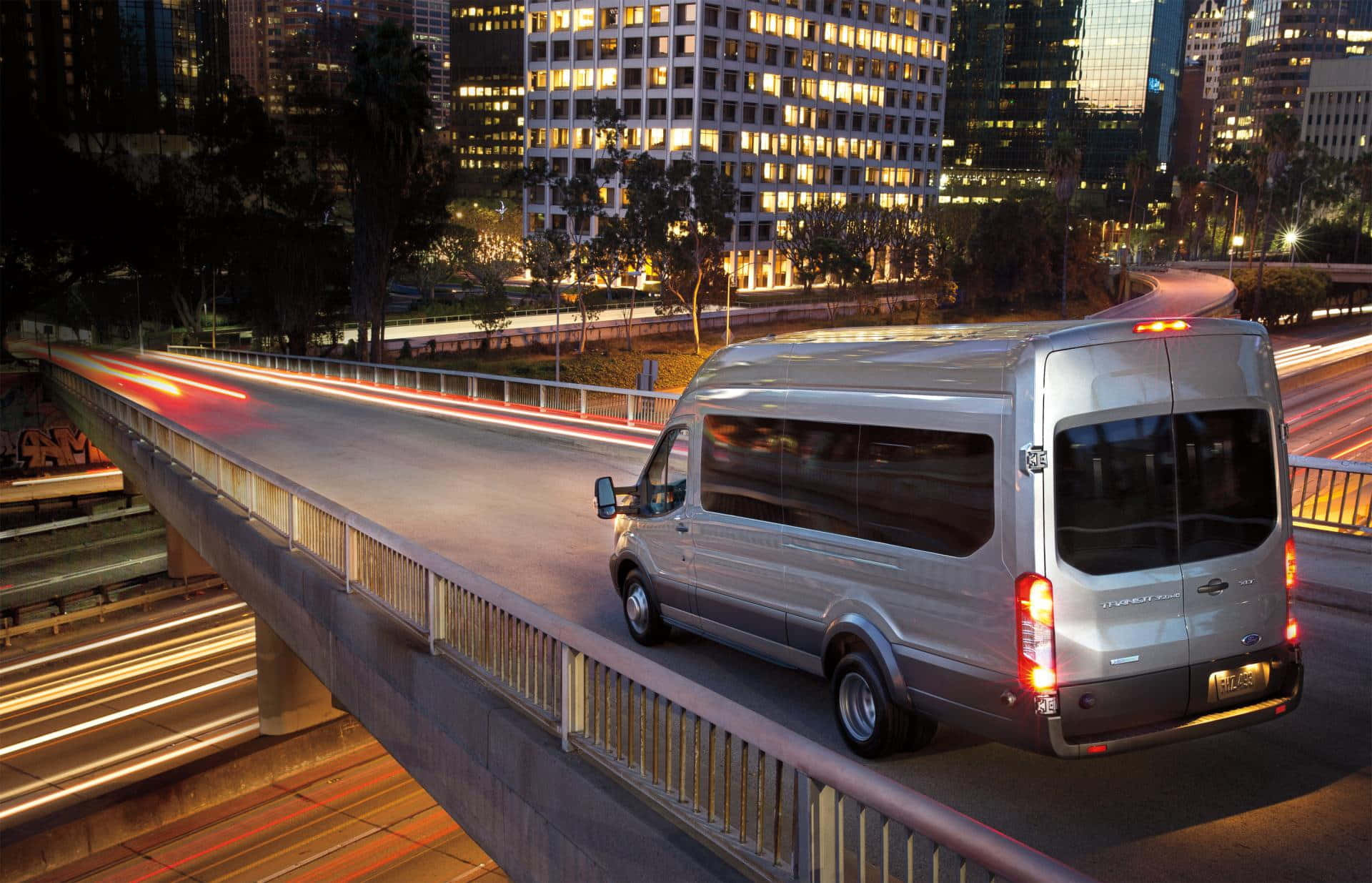 Ford Transit 桌布 壁紙 — 一輛銀色廂型車在夜間行駛於高架高速公路，背景是流動的城市燈光和燈火通明的摩天大樓。