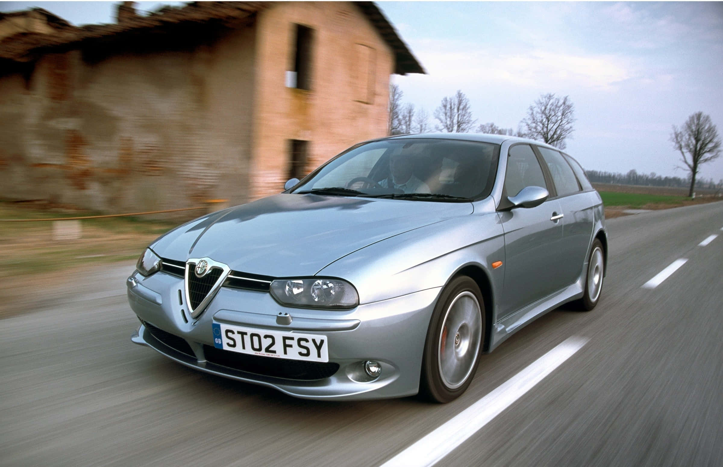 Alfa Romeo 156 桌布 壁紙 — 一輛銀色愛快羅密歐156掀背車在鄉村道路上疾馳，帶有動態模糊效果，背景是一座風化的石頭建築。