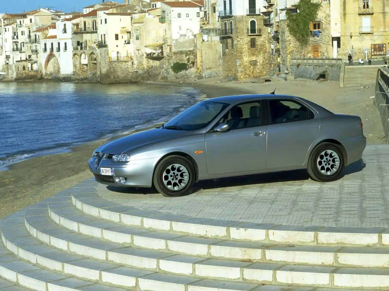 Alfa Romeo 156 桌布 壁紙 — 一輛銀色愛快羅密歐房車停在彎曲的石階上，旁邊是陽光普照的海邊小鎮，有陶土屋頂和寧靜的藍色海水。