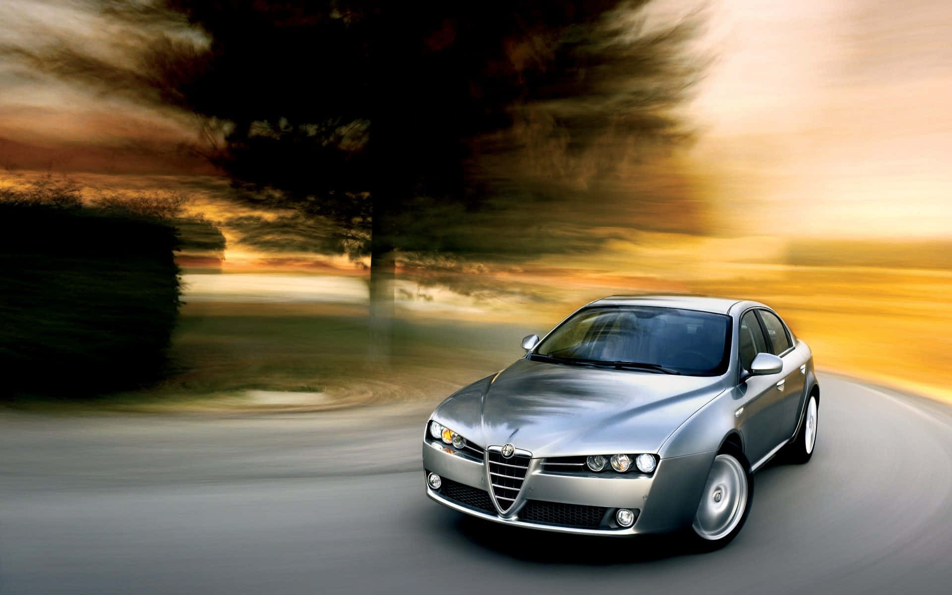 Alfa Romeo 159 桌布 壁紙 — 一輛銀色愛快羅密歐房車在日落時分沿著蜿蜒的道路疾馳，背景是模糊的金色樹木和流動的天空。