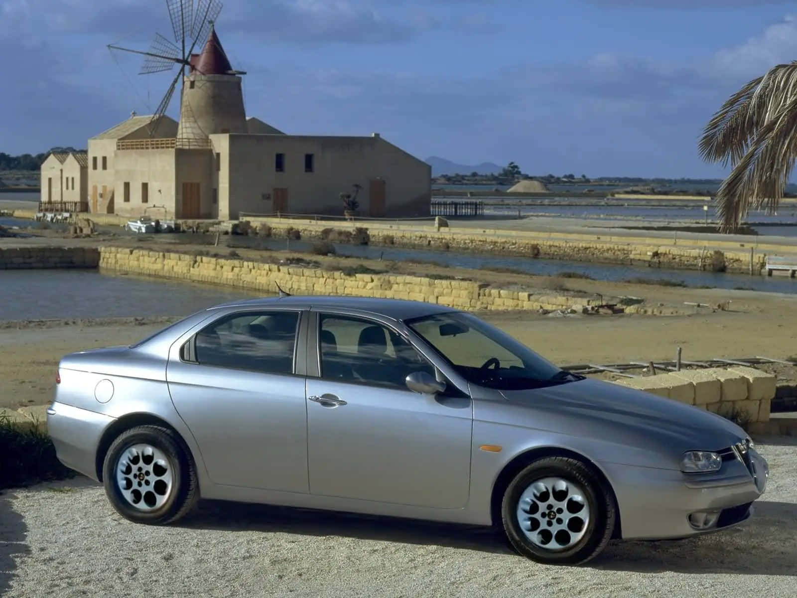 Alfa Romeo 156 桌布 壁紙 — 一輛銀色愛快羅密歐轎車停在碎石路上，靠近歷史風車和鹽田，天空湛藍。