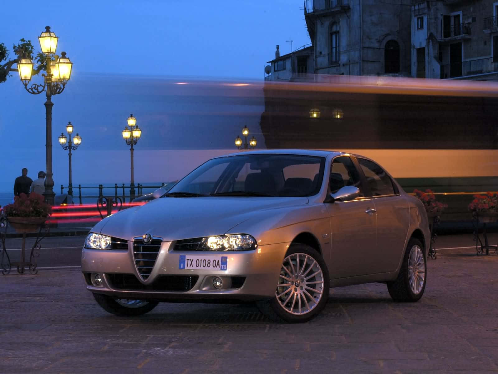 Alfa Romeo 156 桌布 壁紙 — 一輛銀色愛快羅密歐轎車在暮色街燈下閃耀，一輛模糊的巴士駛過歐洲鵝卵石步道。