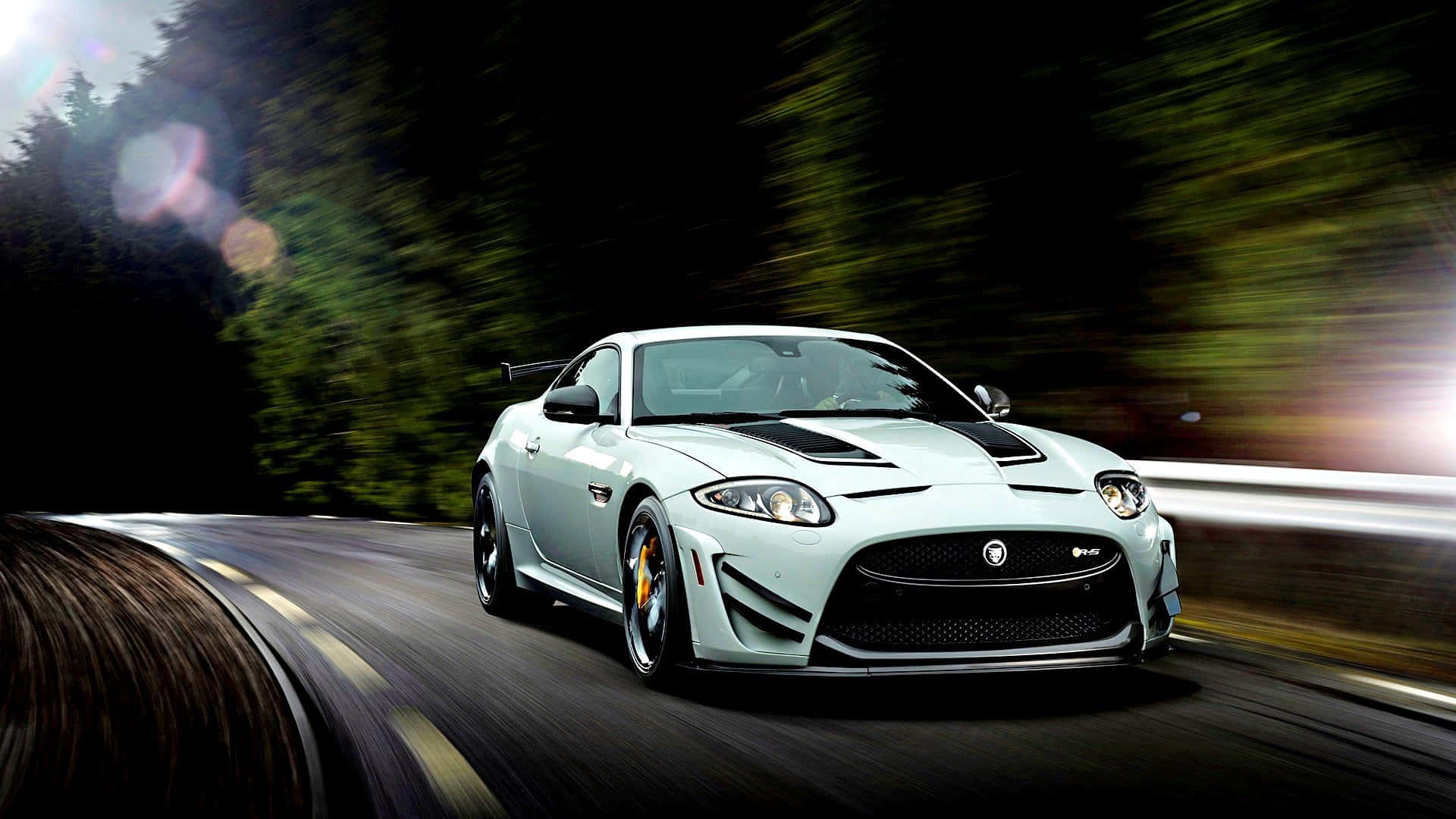 Jaguar Xkr 桌布 壁紙 — 一輛銀色捷豹跑車在蜿蜒的森林道路上疾馳，帶有動態模糊和鏡頭光暈。
