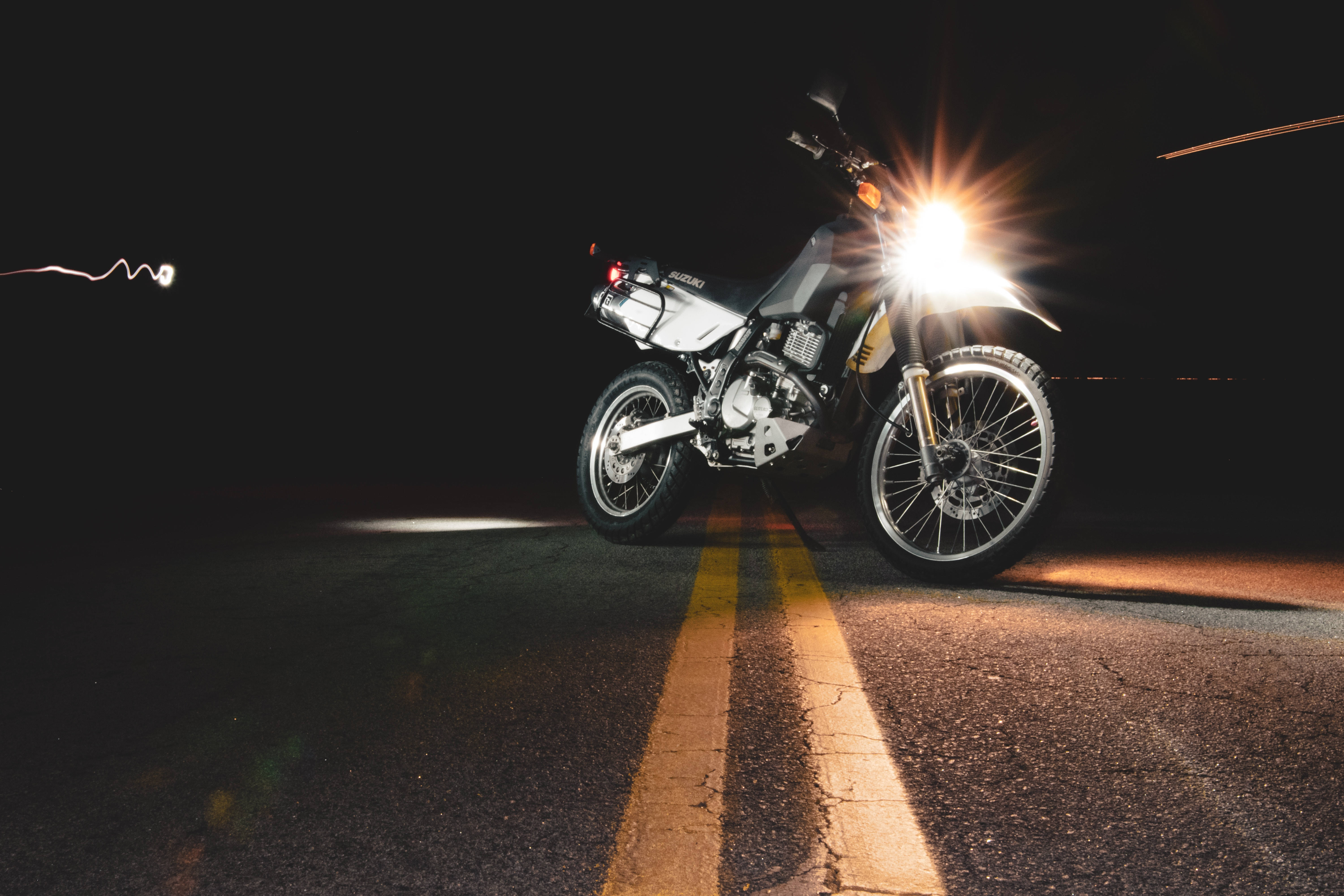機車 桌布 壁紙 — 一輛銀色摩托車在夜間亮著發光的頭燈，停在黑暗的道路上，照亮了破裂的瀝青和黃線。