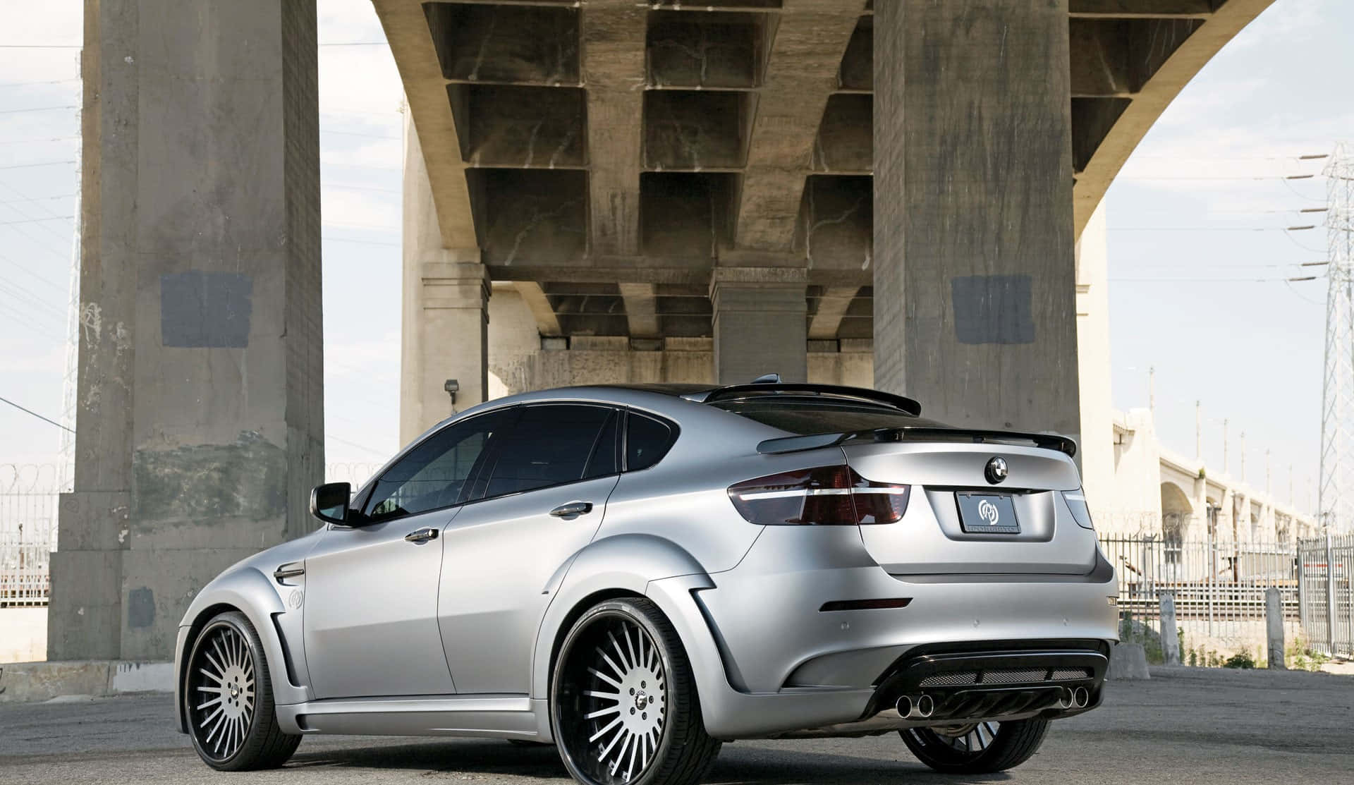 Bmw X6 M 桌布 壁紙 — 一輛銀色改裝BMW M6雙門跑車停放在混凝土高架橋下，背景有工業圍欄和電線。