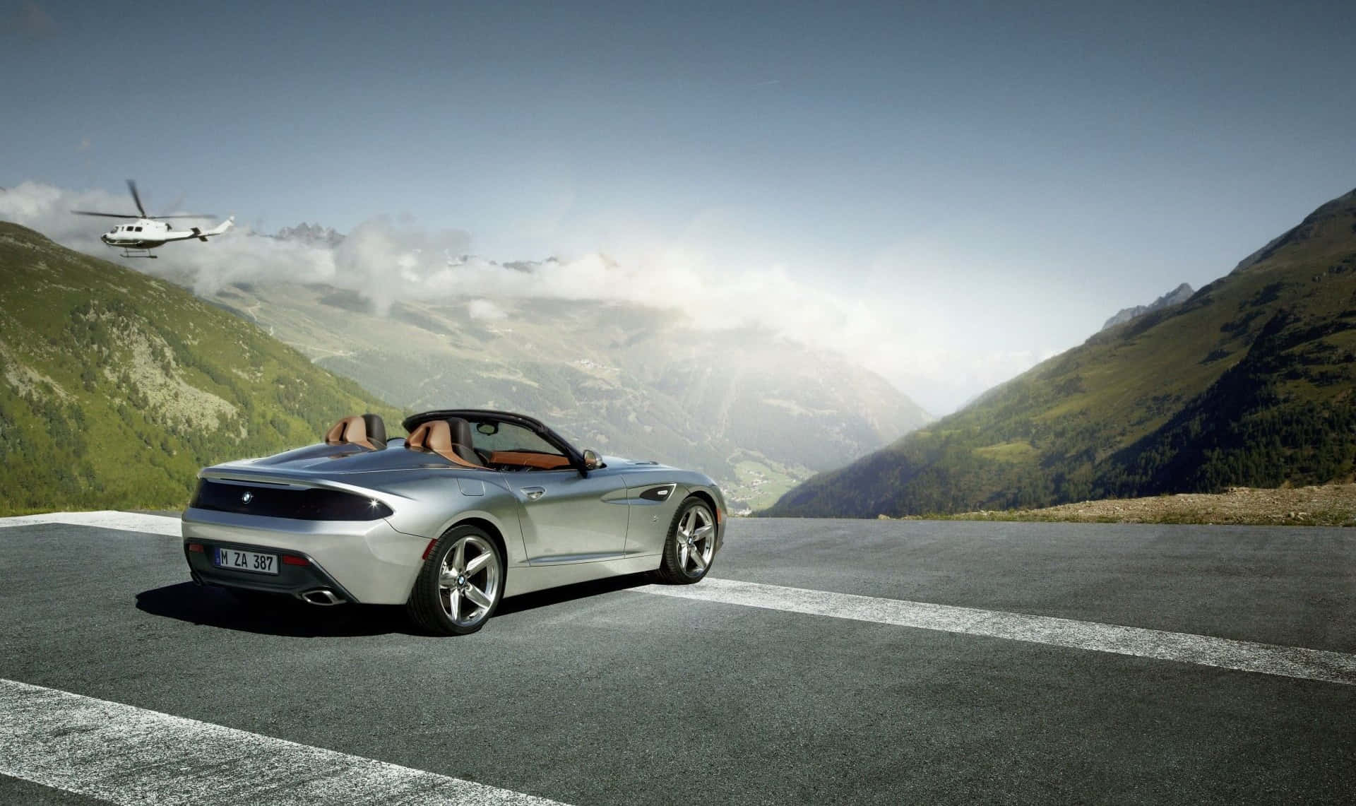 Bmw Z4 桌布 壁紙 — 一輛銀色敞篷跑車停在山路上，一架直升機在頭頂飛過，背景是綠色的山峰和雲層。