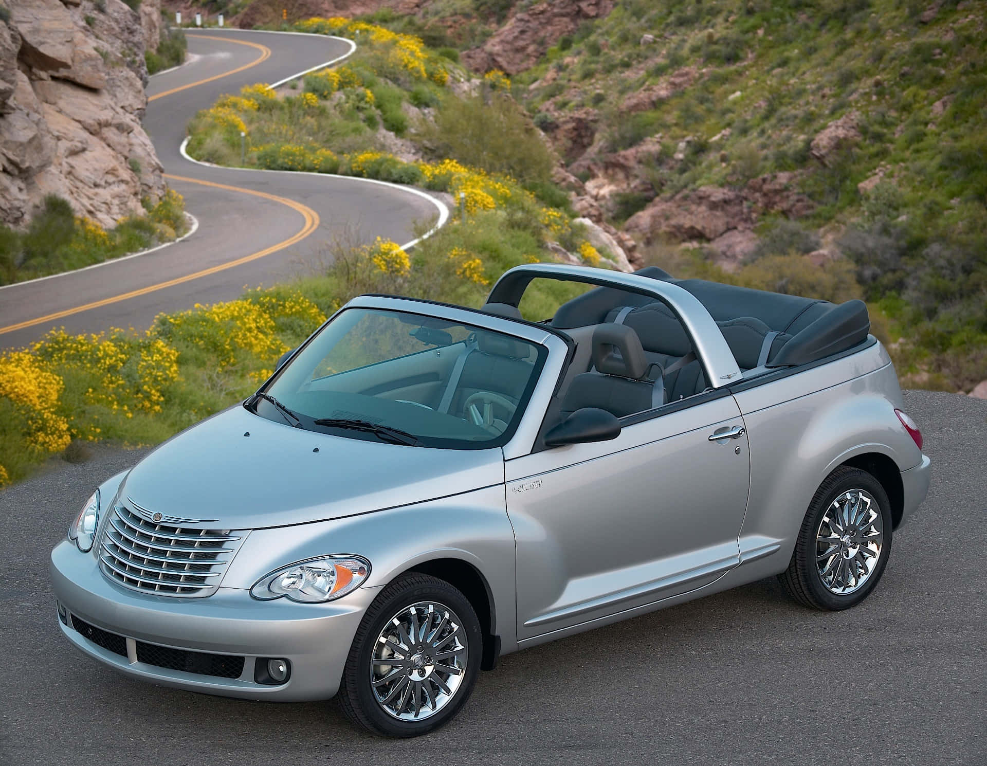 Chrysler Pt Cruiser 桌布 壁紙 — 一輛銀色敞篷跑車停放在蜿蜒的山路上，周圍環繞著黃色野花和岩石地形。