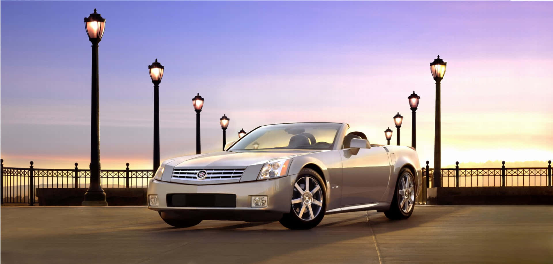 Cadillac Xlr 桌布 壁紙 — 一輛銀色敞篷跑車停泊在日落時分的碼頭上，發光的燈柱排列在步道旁。